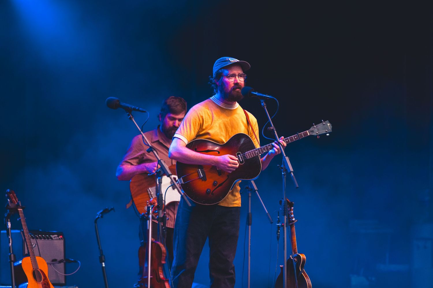 Bearded man in yellow shirt playing guitar at a microphone on a blue-lit smoky stage.