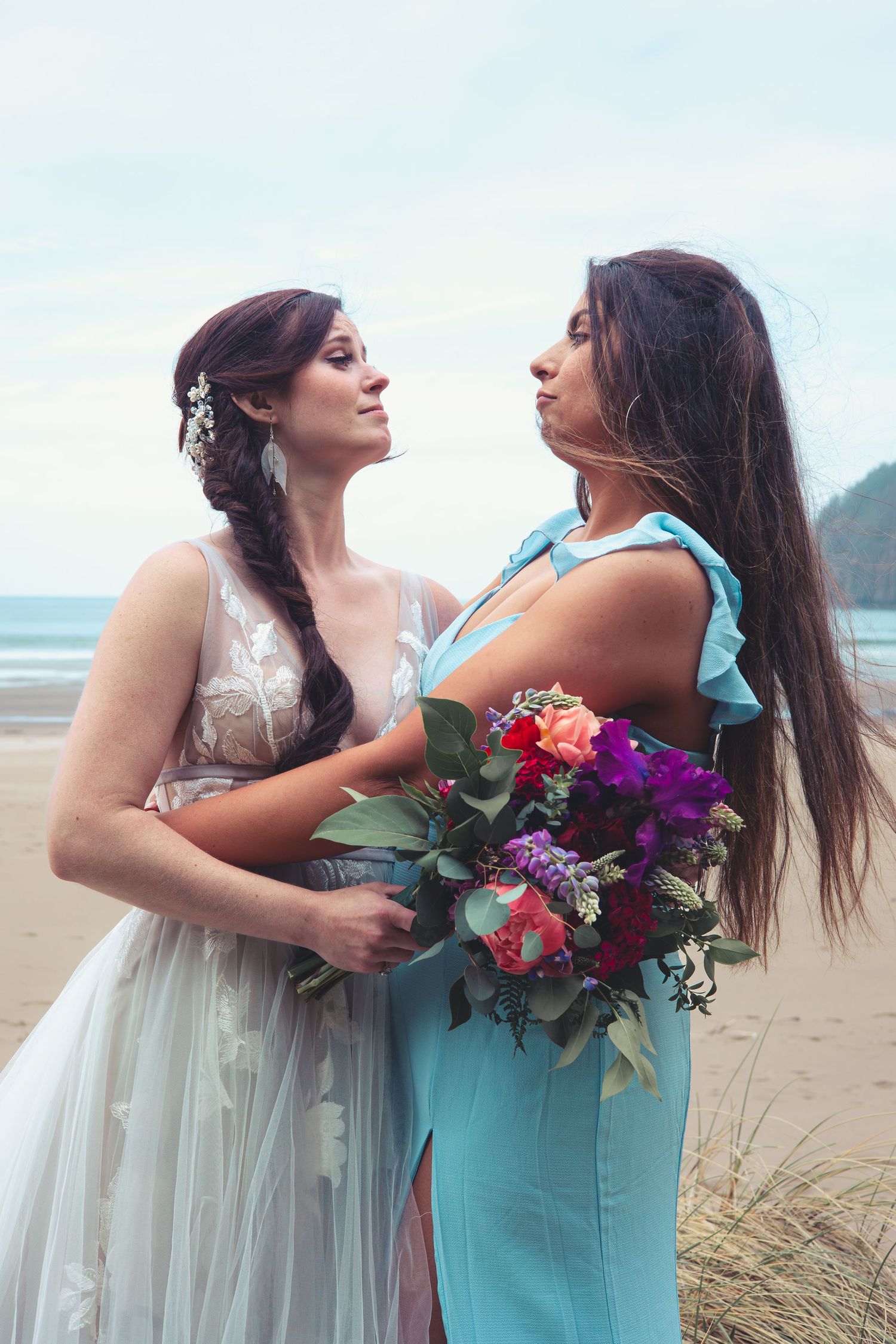 Two women in elegant wedding attire stand together on a sandy beach, holding a vibrant floral bouquet.
