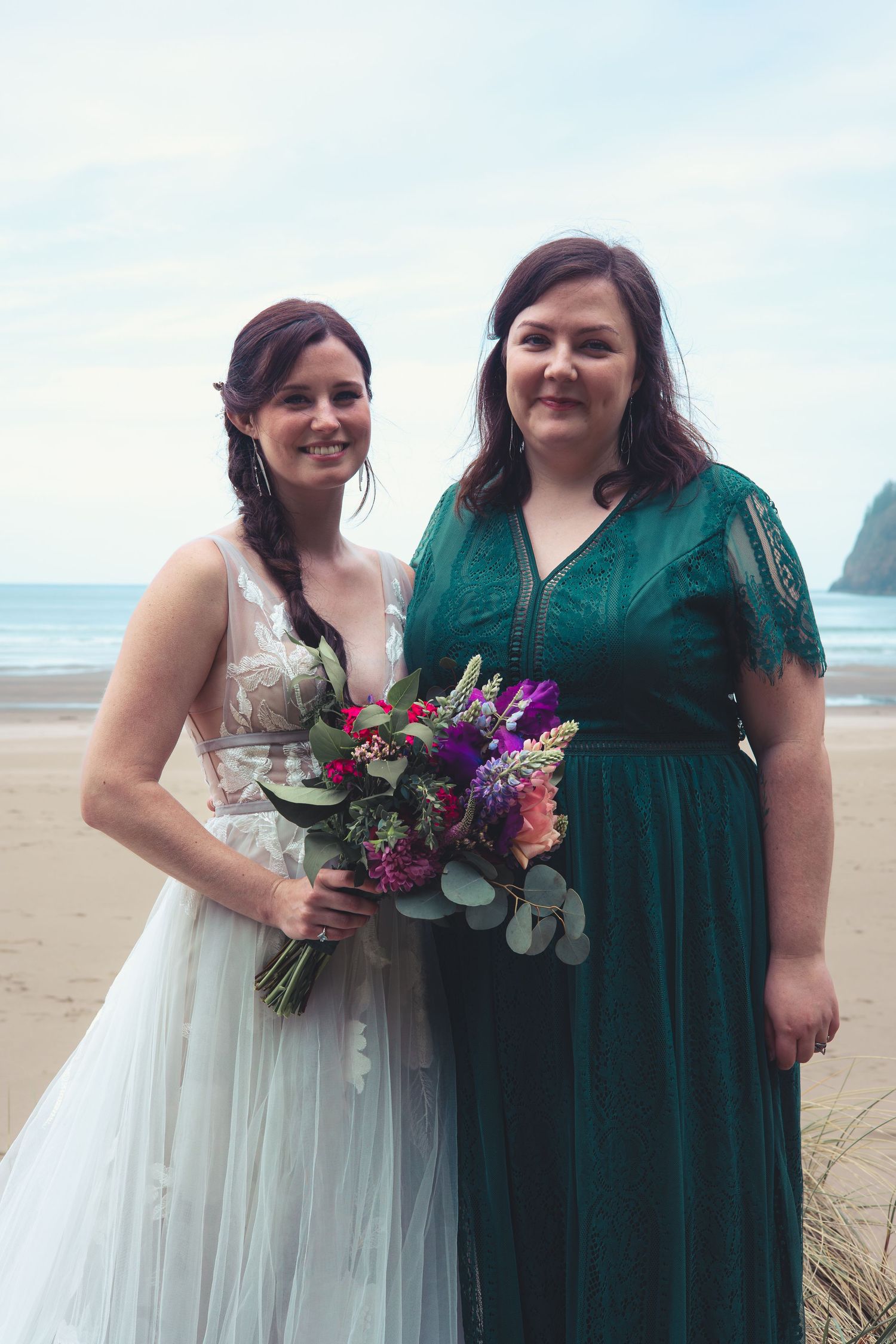 A bride in a white wedding gown and bridesmaid in a dark green dress smile on a sandy beach.