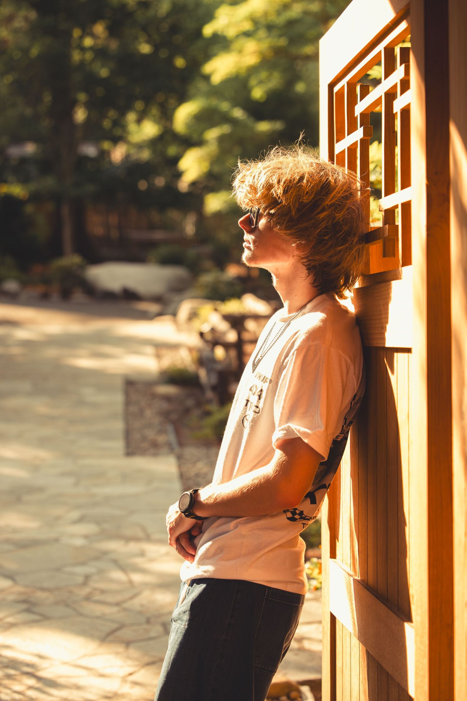 Young man with curly hair and sunglasses leaning against a wooden wall in golden sunlight outdoors.