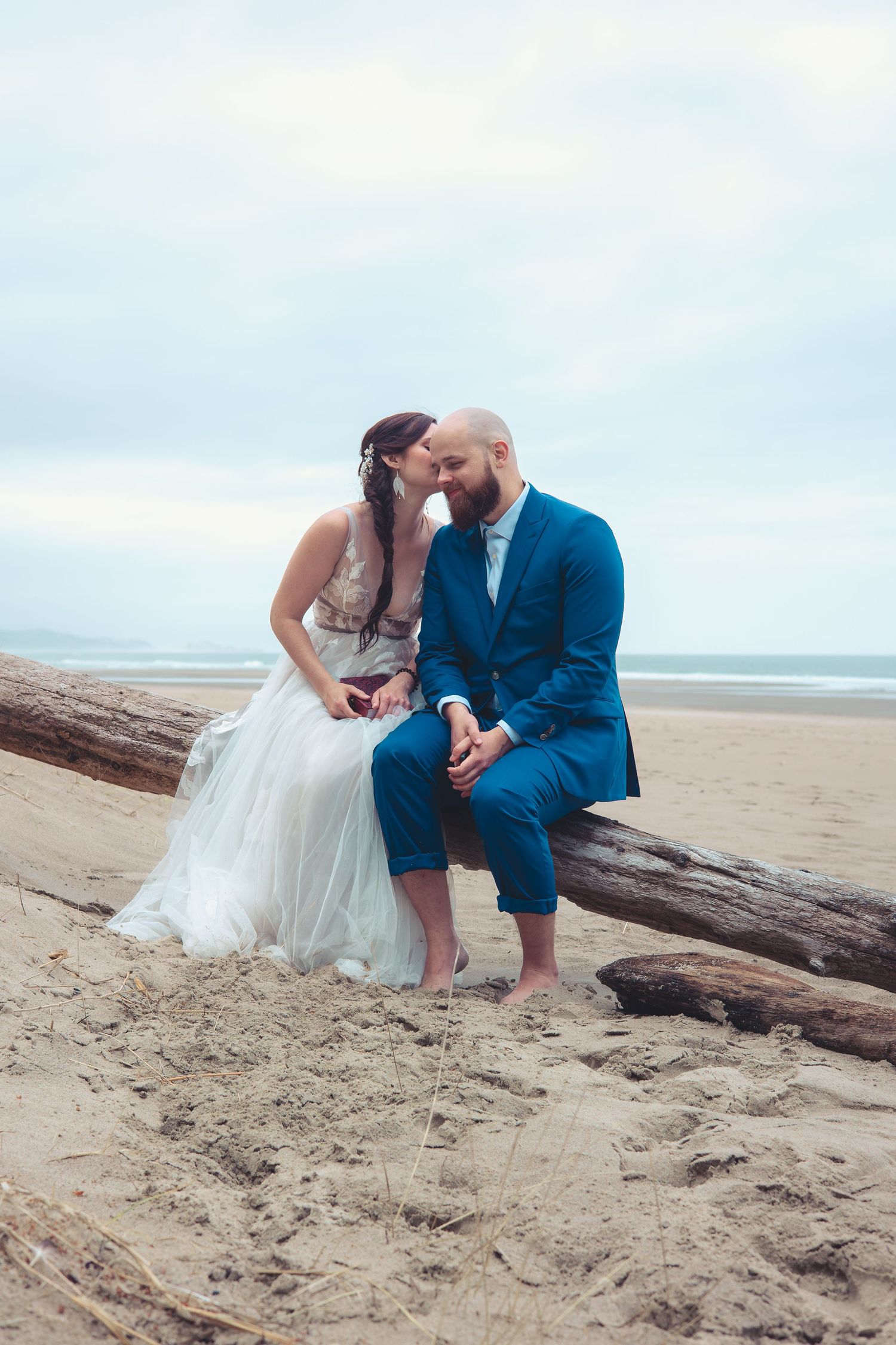 Bride leans in close to groom as they sit together on a driftwood log on a serene sandy beach.