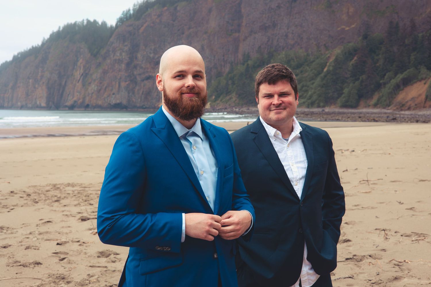 Two men in blue suits smiling on a sandy beach with rocky cliffs in the background.