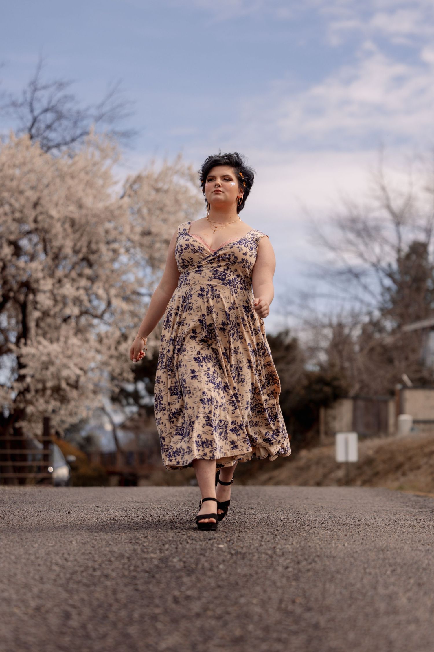 Plus-size woman in vintage floral sundress strolling down rural road with blossoming trees nearby.