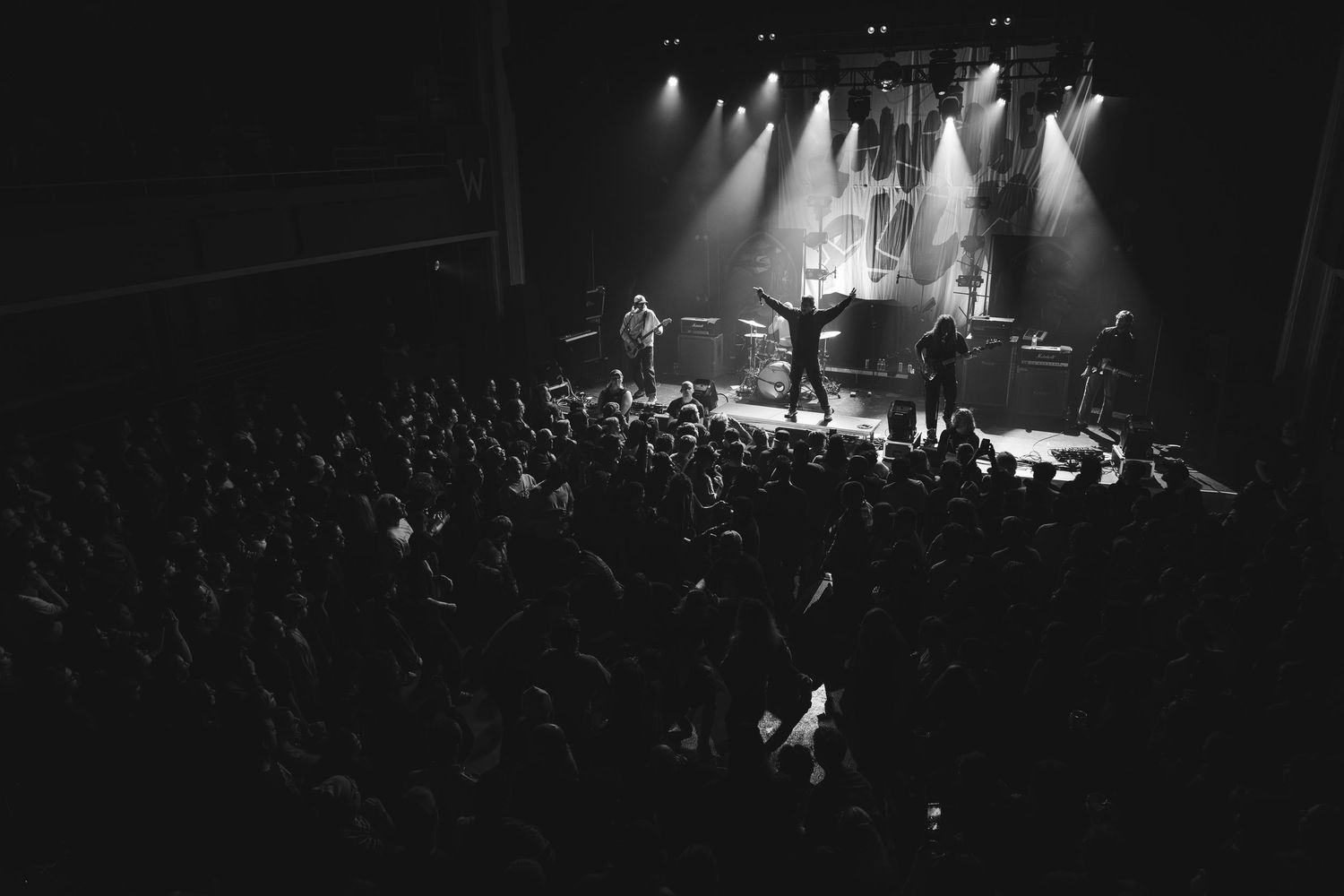 Black and white wide-angle shot of band performing on lit stage before massive crowd at indoor venue.