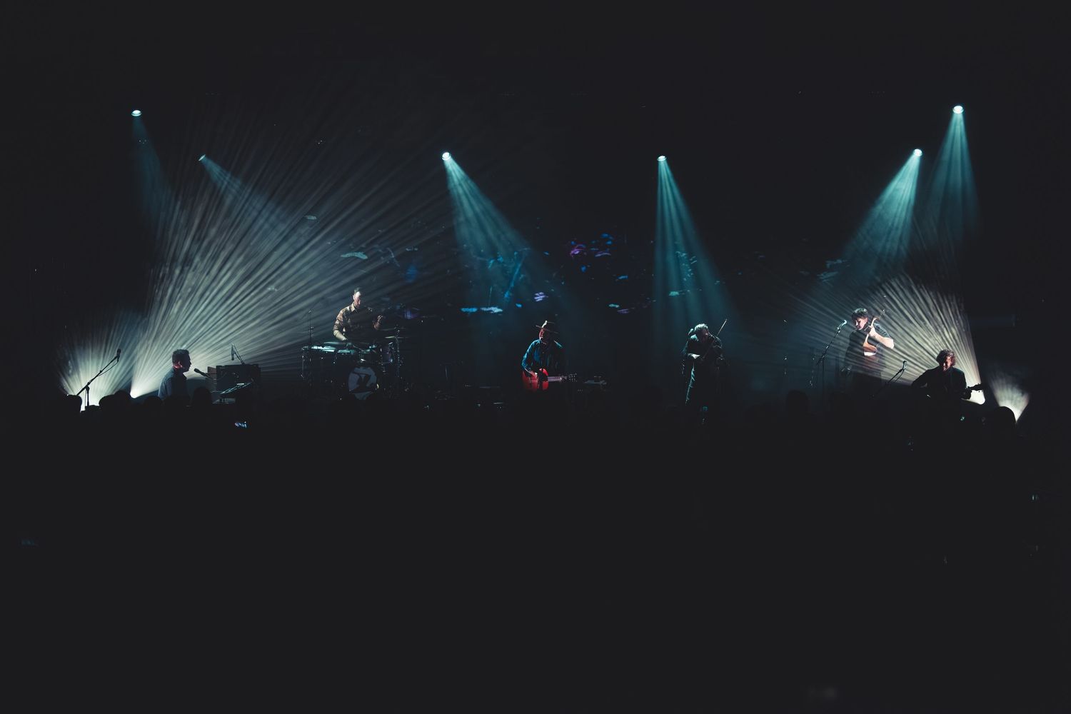 Band performing on dark stage illuminated by dramatic white laser light beams radiating outward in concert venue.