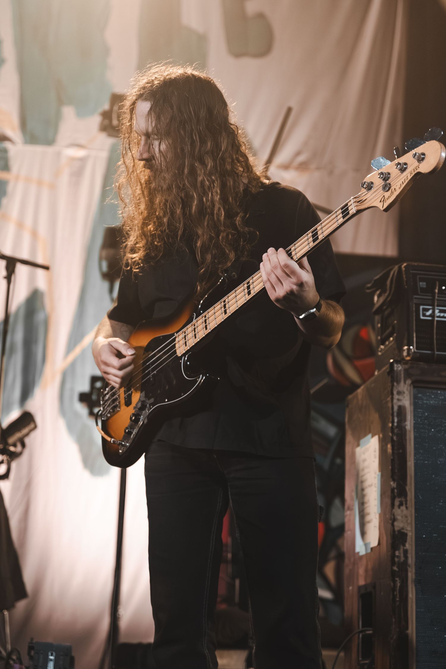 Long-haired bassist playing a sunburst electric bass guitar on stage during a live music performance.