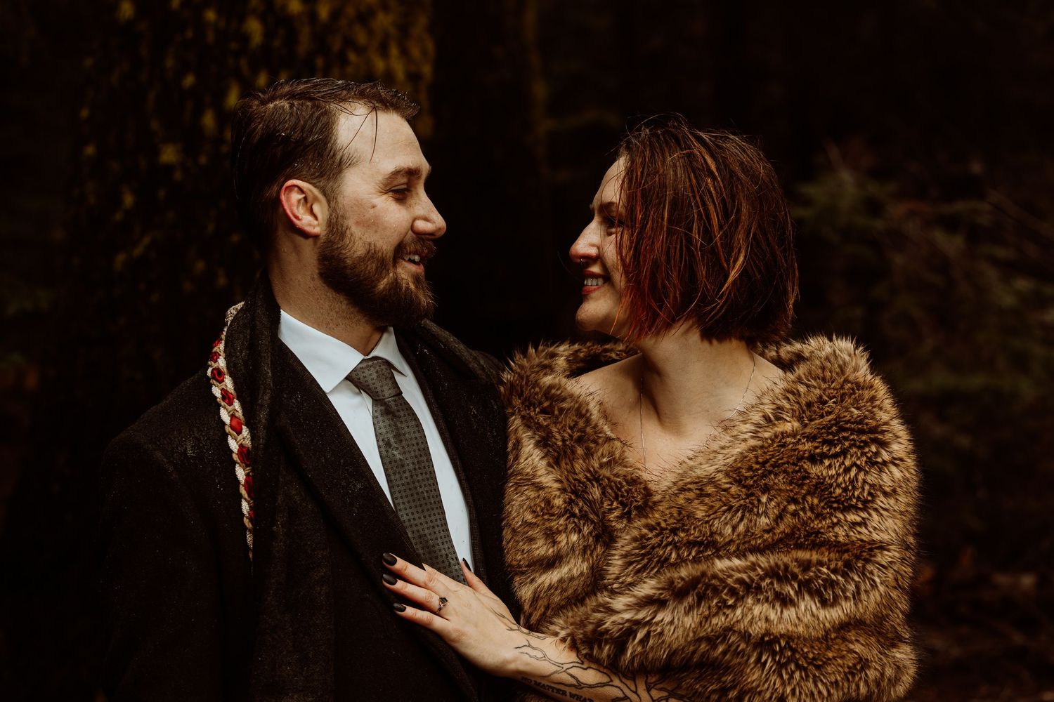 Bride in fur stole and groom in dark suit smiling at each other in an autumn forest wedding portrait.