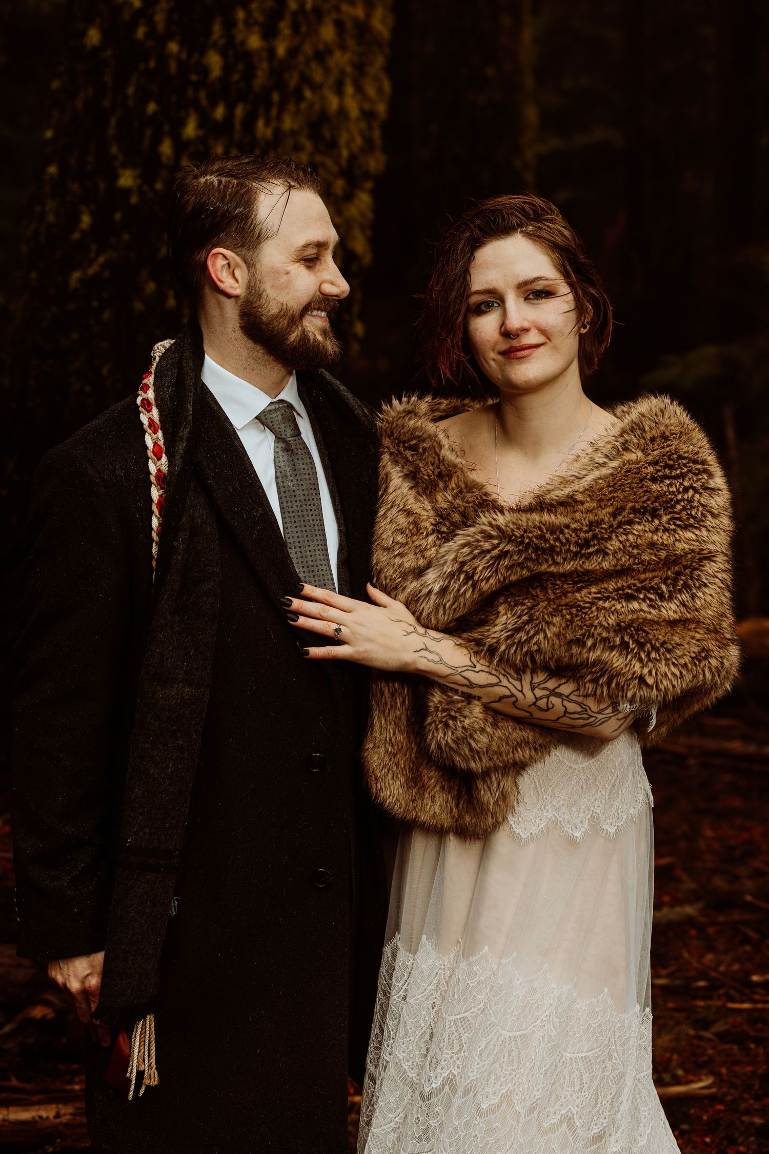 Bride in fur wrap and groom in dark coat sharing a tender moment in a moody forest setting.
