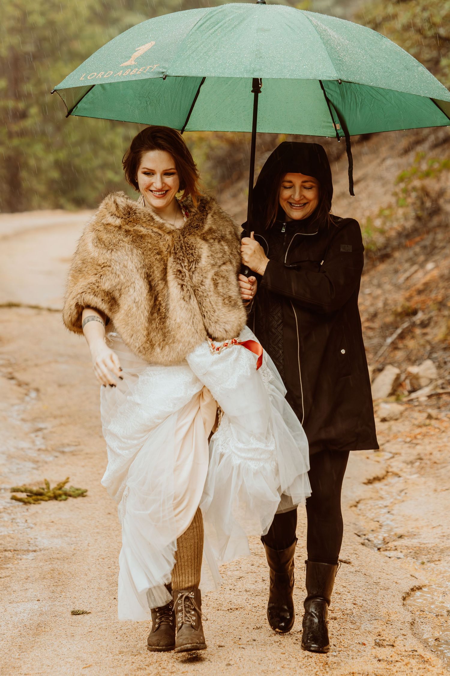 Bride in fur stole lifts wedding dress while walking in rain with woman holding green umbrella on dirt path.