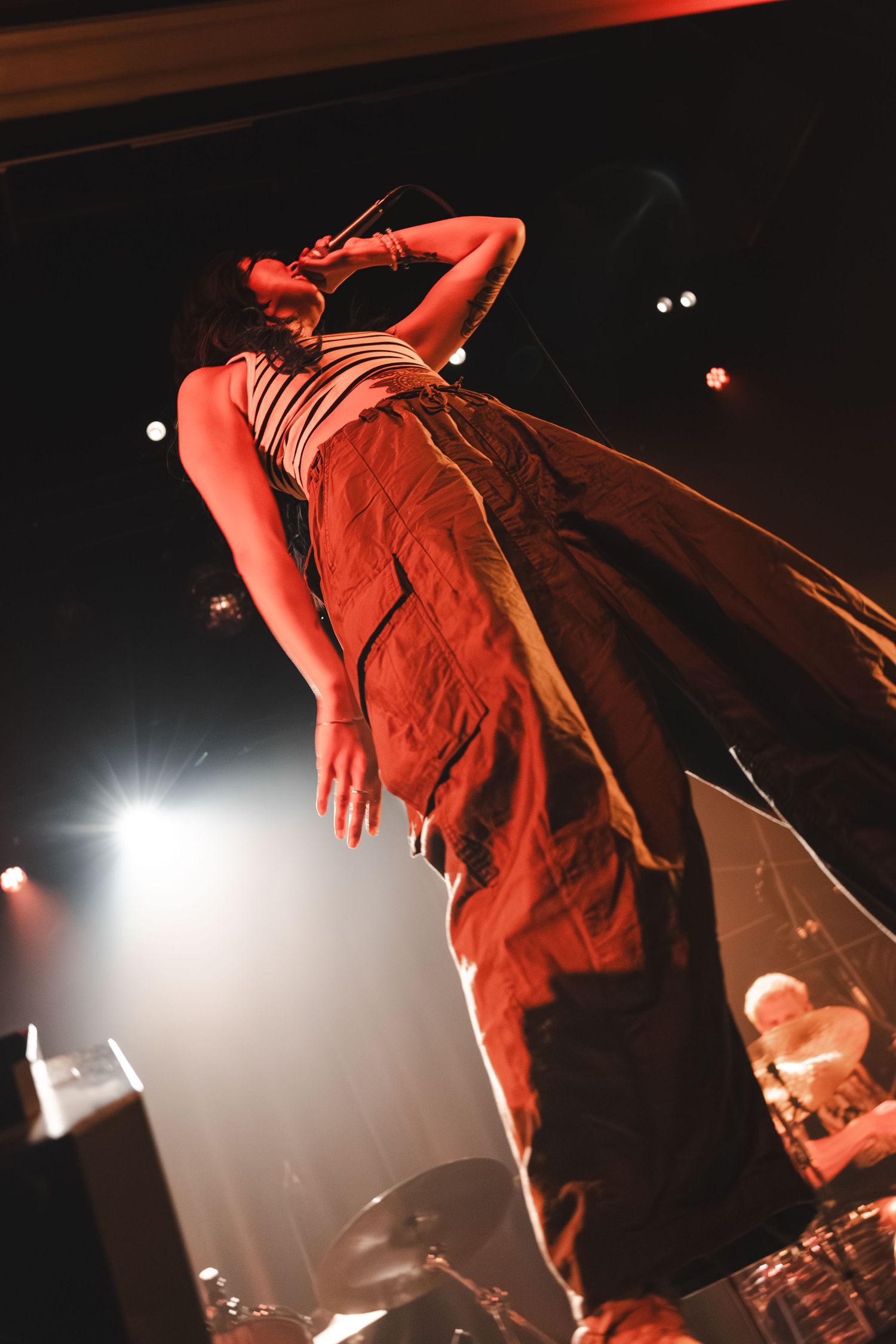 Low angle concert shot of vocalist leaning back dramatically on stage under warm red and white stage lights.