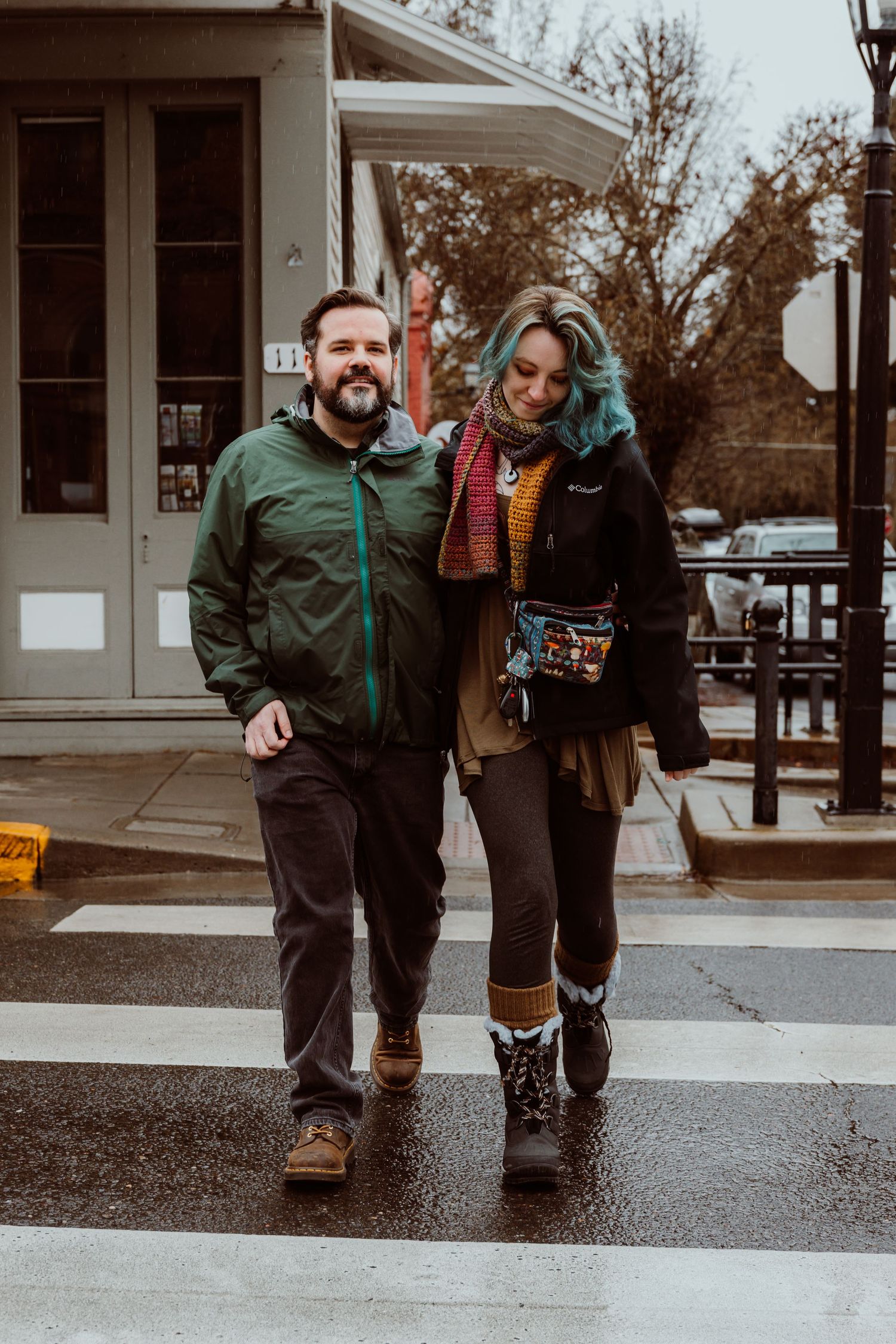 A smiling bearded man and blue-haired woman crossing a wet street together.