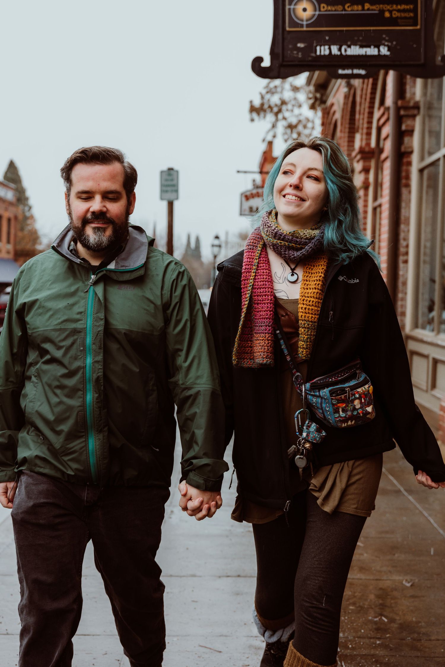 A smiling couple holding hands on a sidewalk, woman with teal hair wearing a colorful scarf.