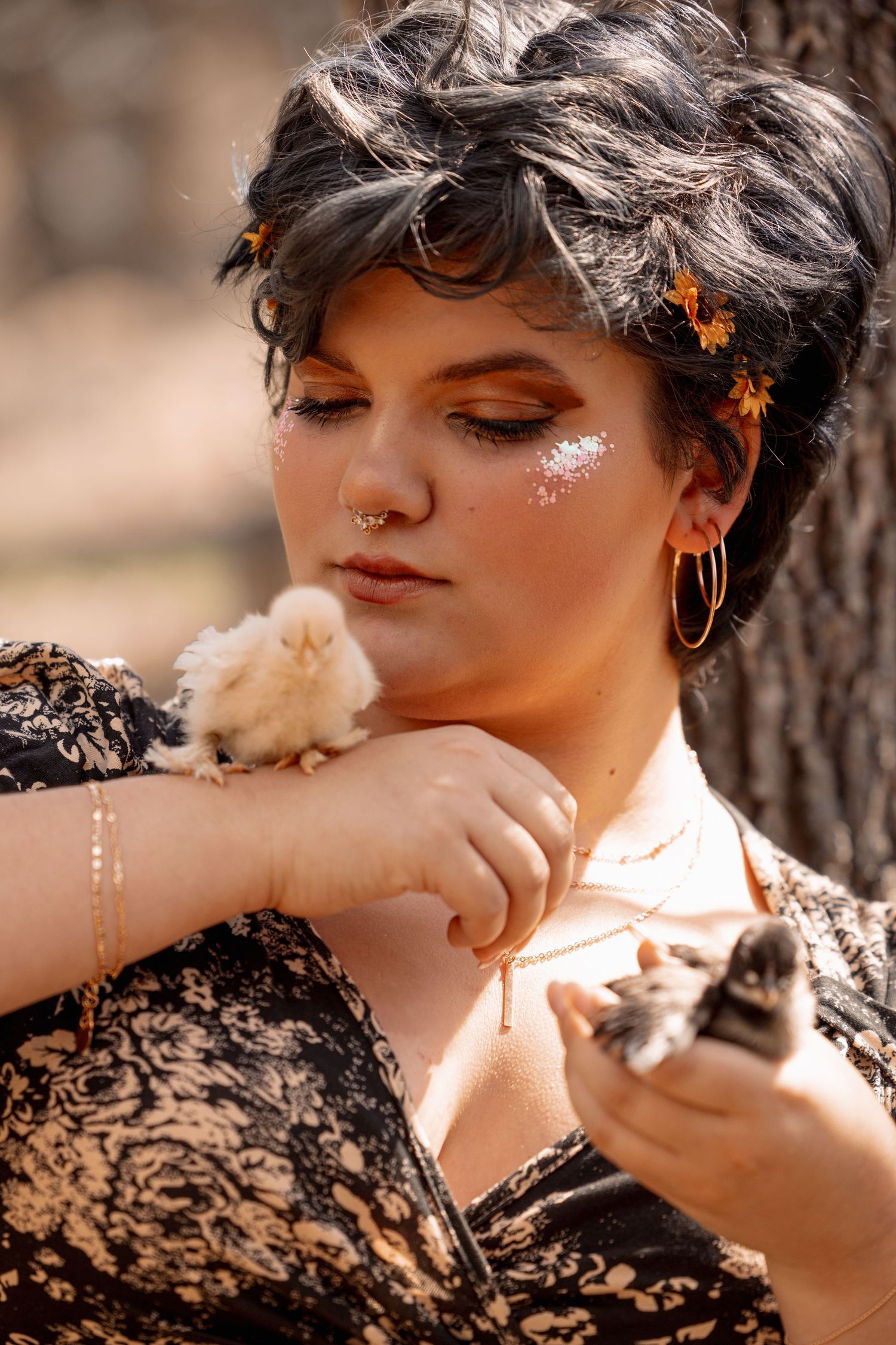 Woman with glitter makeup and hoop earrings tenderly holds two baby chicks against her chest outdoors.