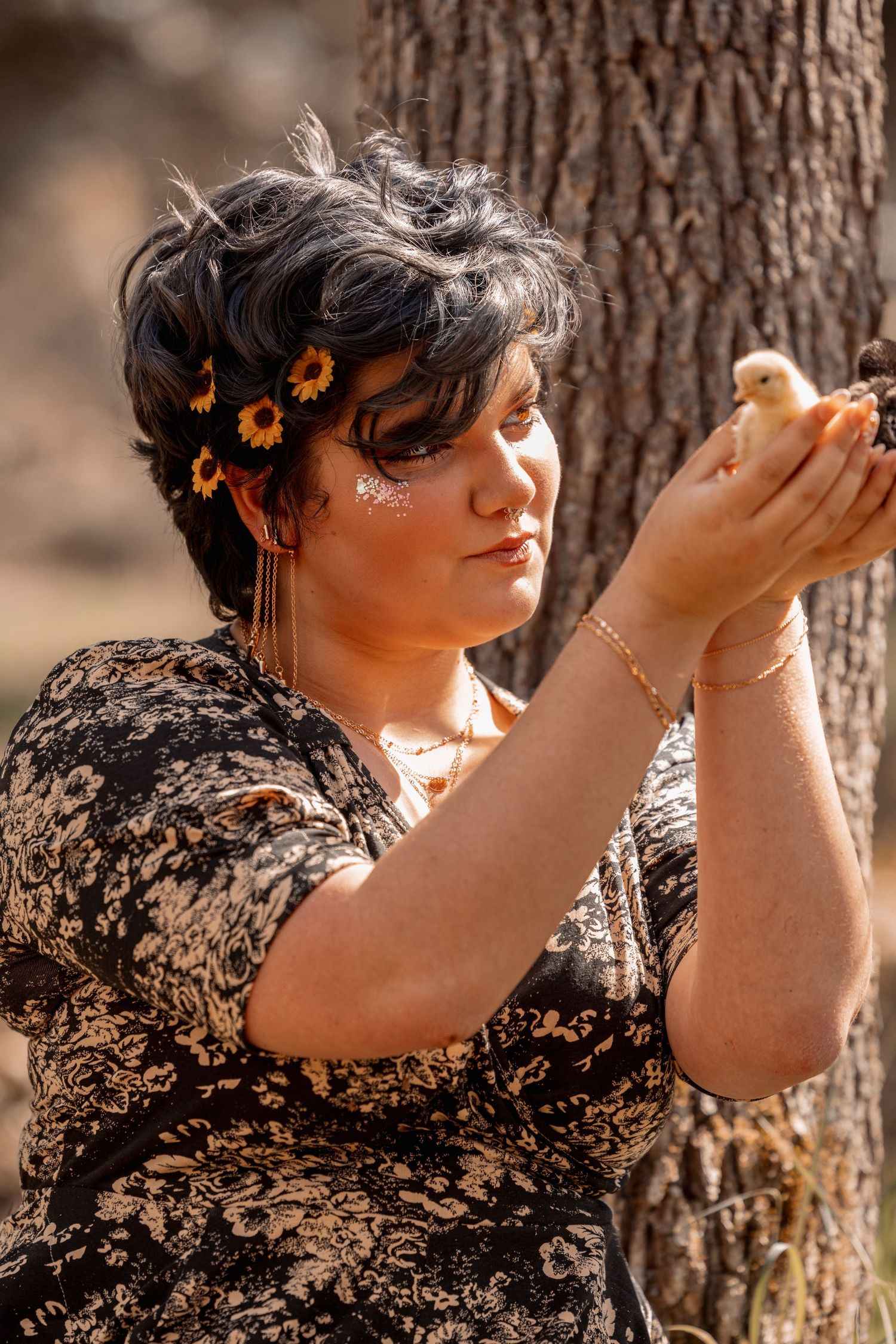 Woman with sunflower hair accessories holds a tiny yellow chick in her hands outdoors near a tree.