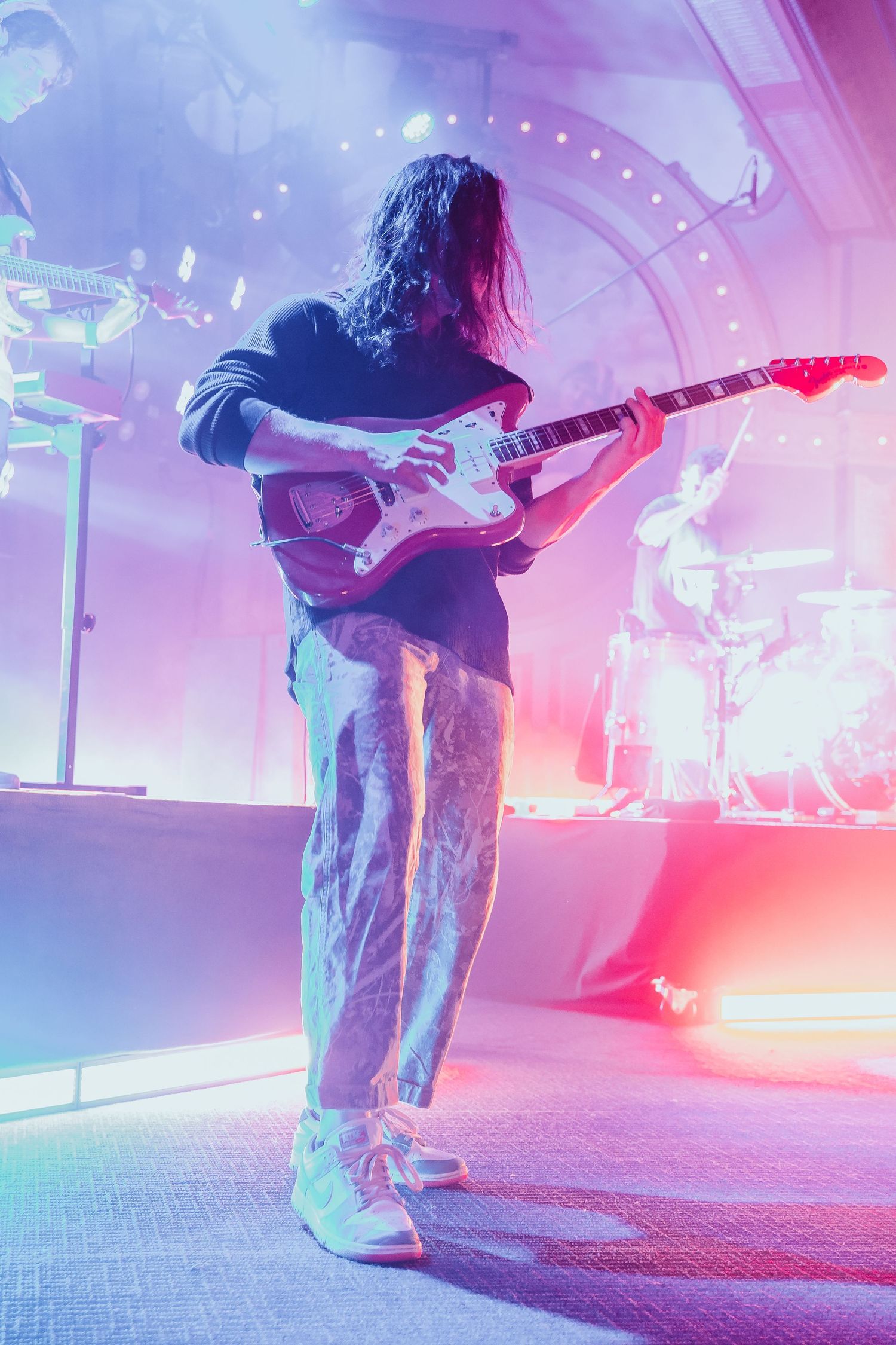 Guitarist with long hair playing red Fender Jazzmaster on a foggy, colorfully lit stage.