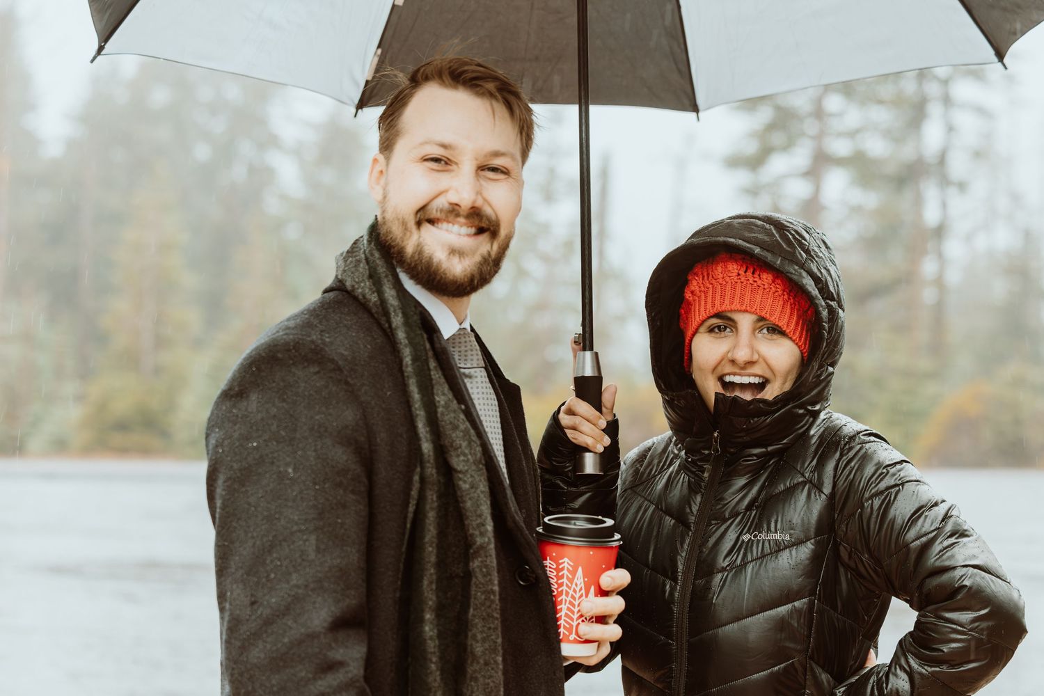 Smiling couple sharing umbrella and holiday coffee cups in rainy outdoor setting.