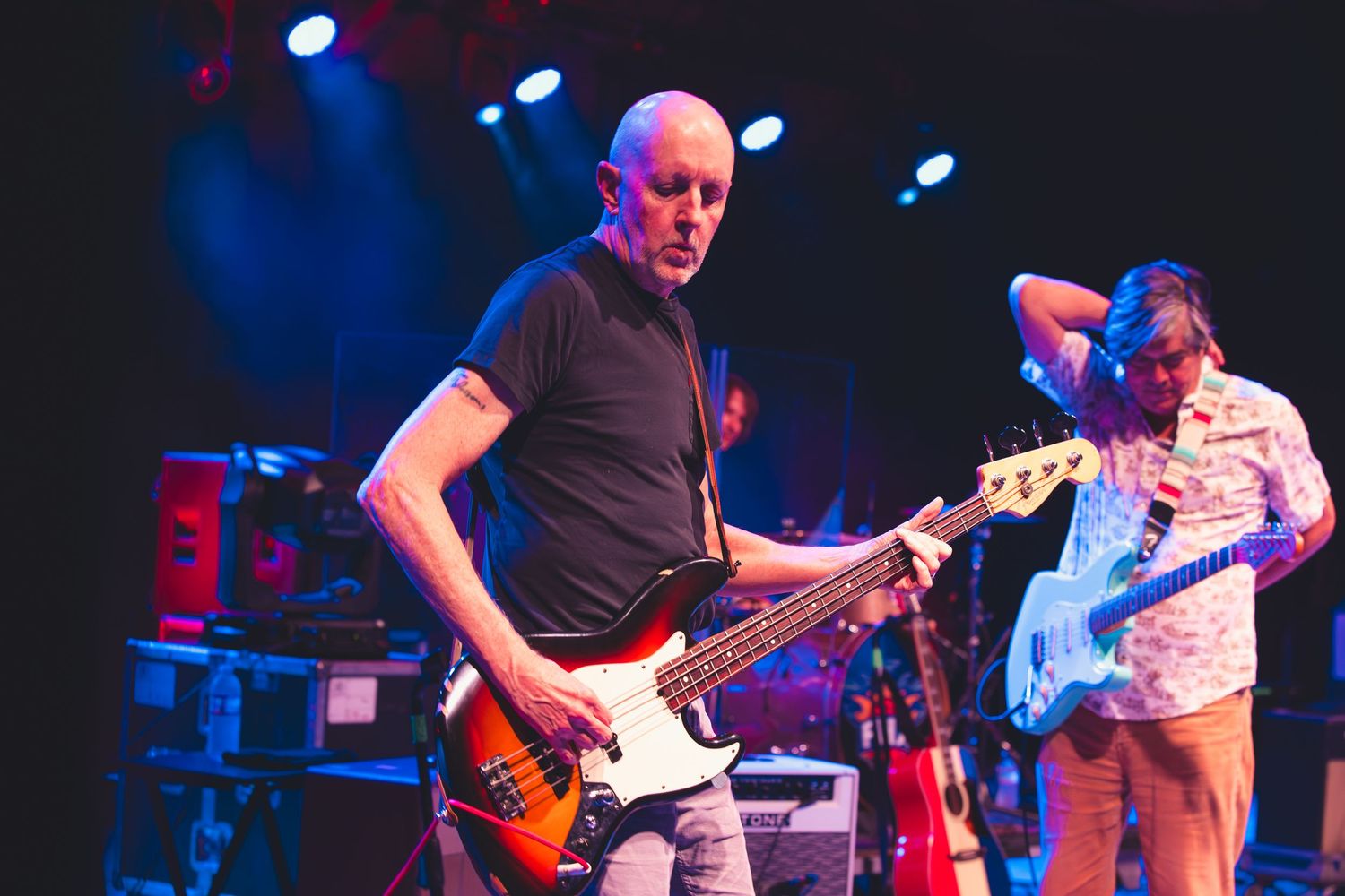 Two guitarists perform on stage, the foreground player holding a sunburst bass guitar.