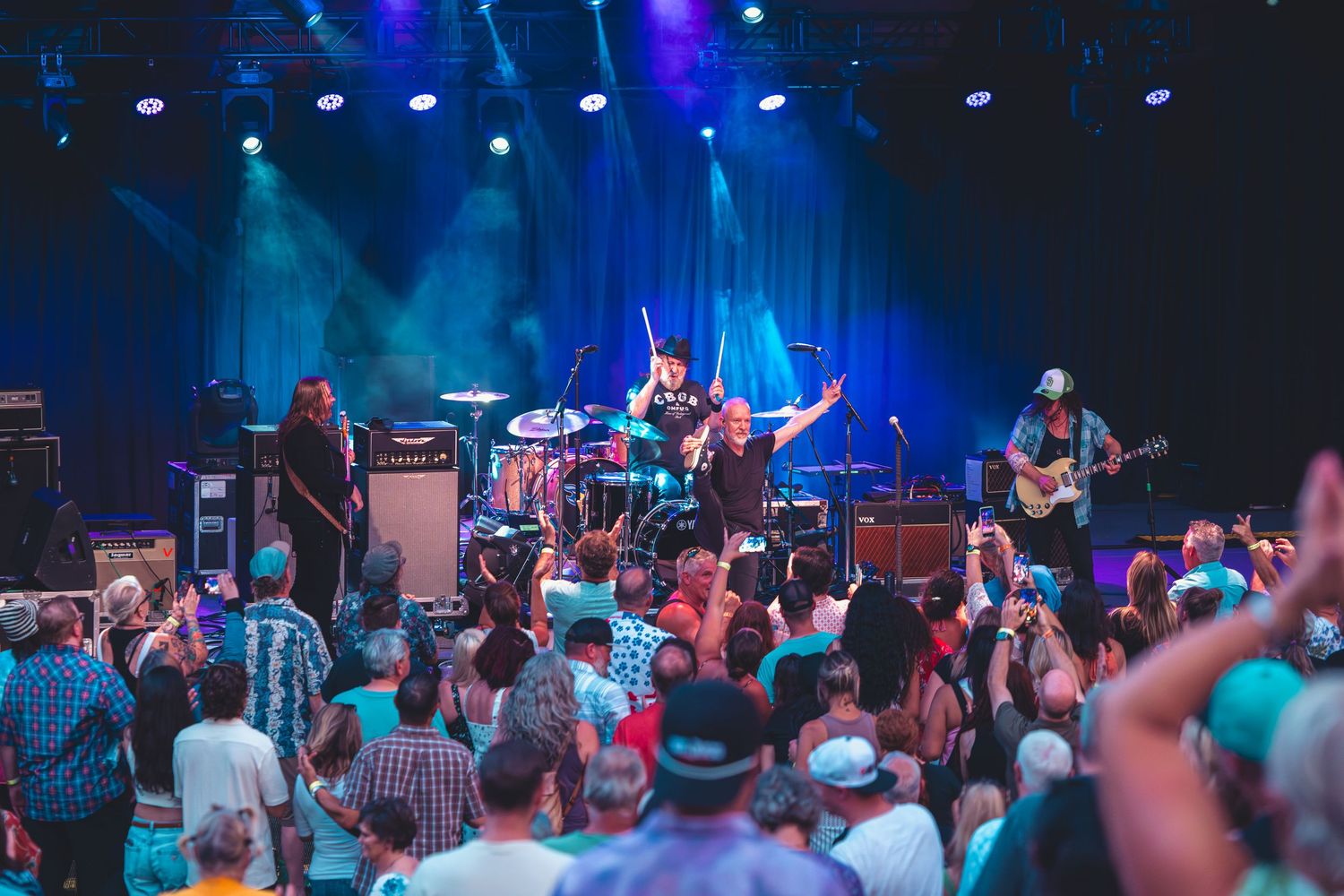 Live rock band performing on stage with colorful lights before a large crowd at an outdoor concert.