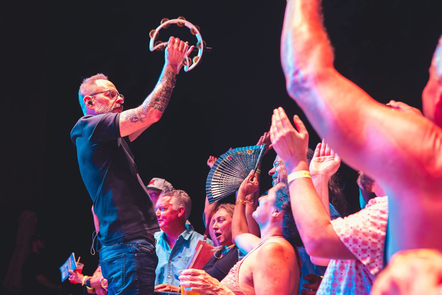 Tattooed performer holding a tambourine above crowd of enthusiastic fans reaching up at a live concert.