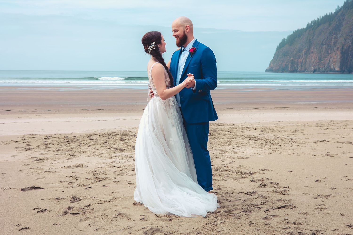 Bride and groom dance barefoot on sandy beach, facing each other in white gown and blue suit, cliffs behind.