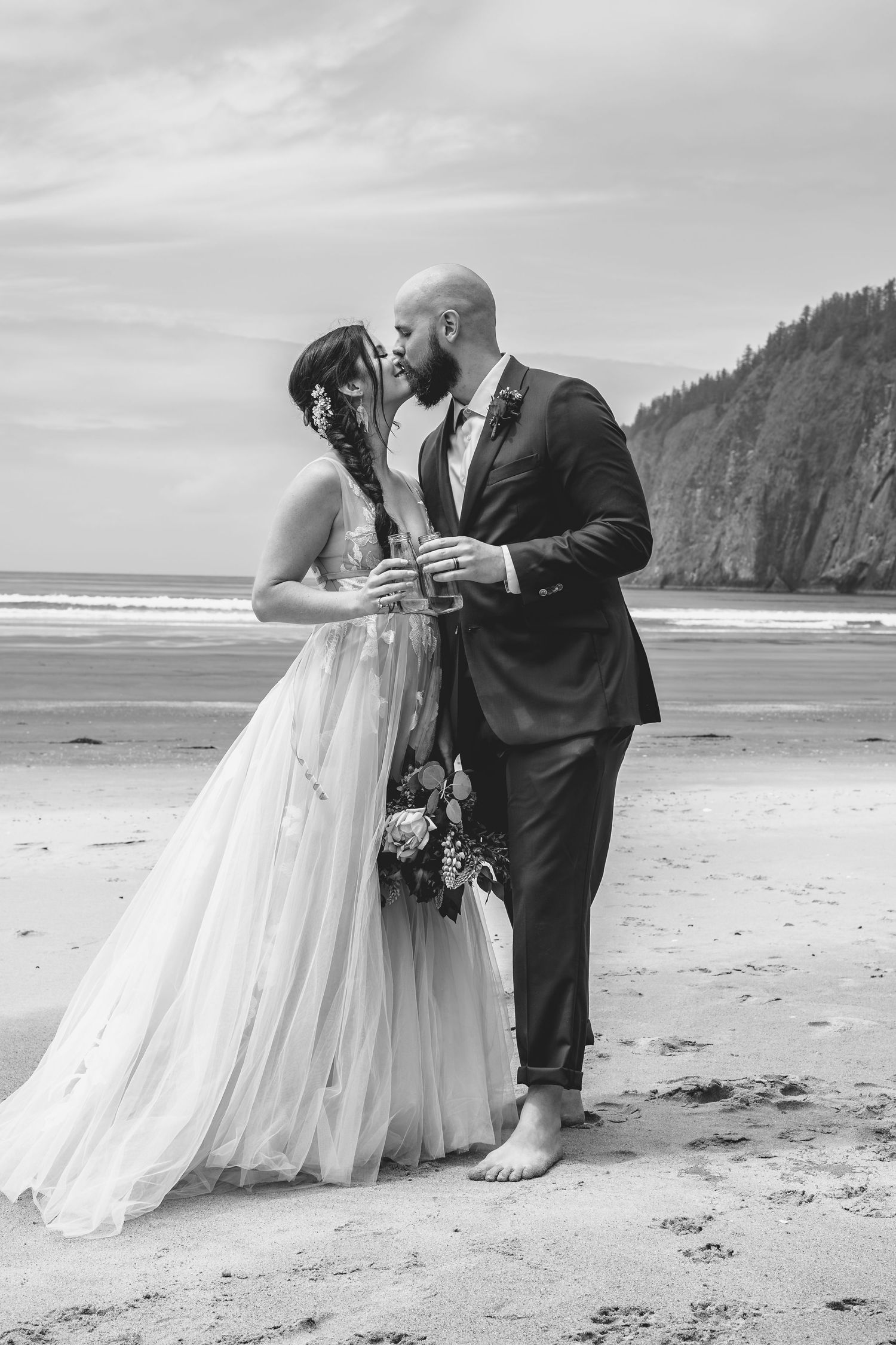 Black and white photo of bride and groom kissing on a beach while holding champagne glasses.