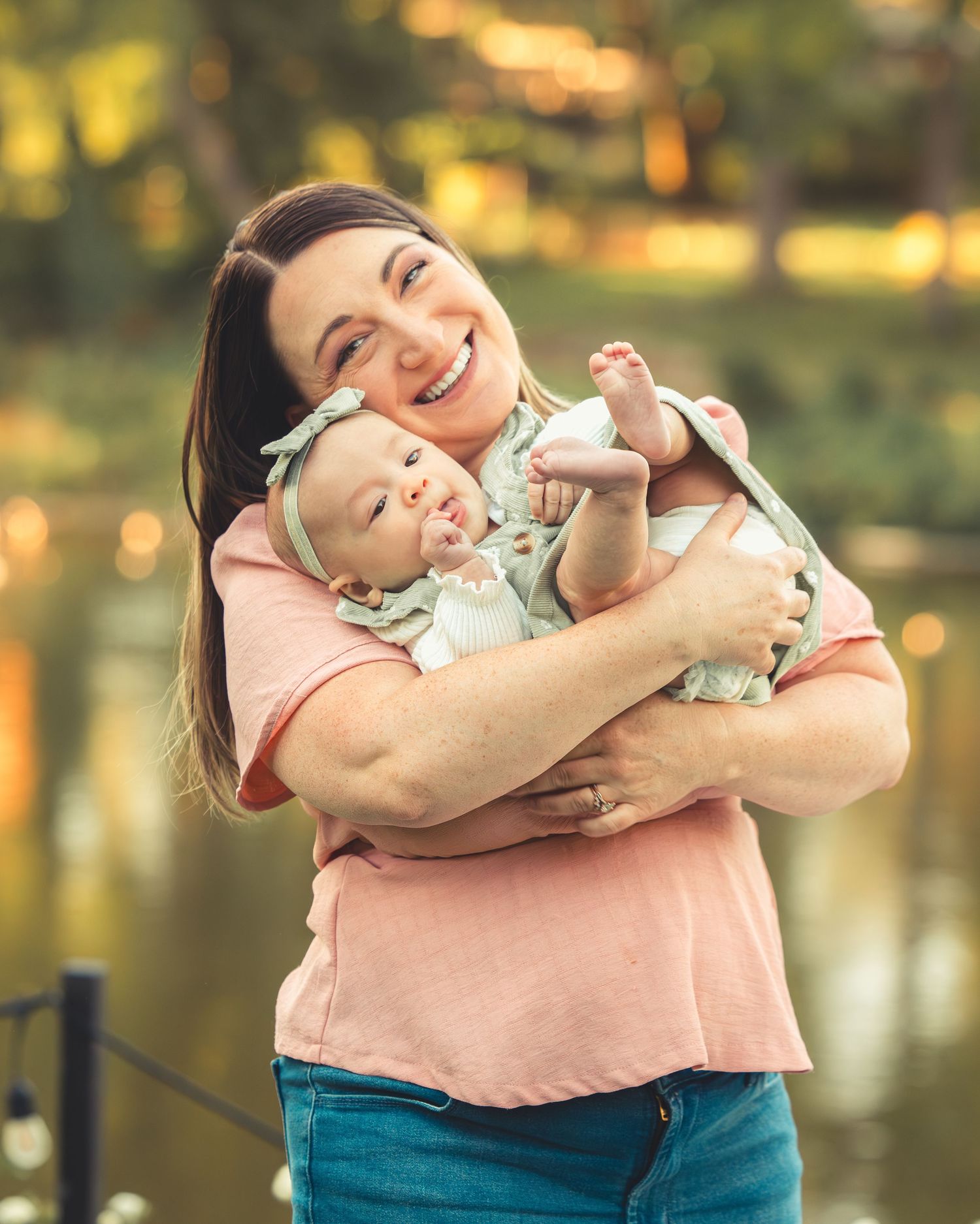 Joyful mother holding her newborn baby girl by a glowing pond in a warm autumn park setting.