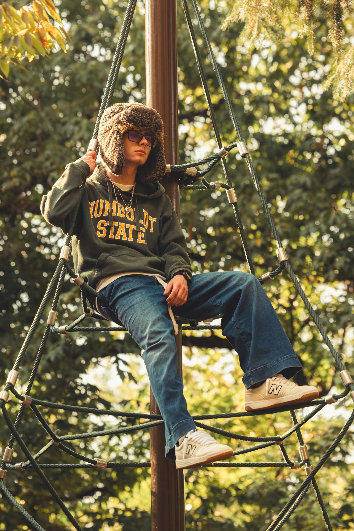 Teen in Humboldt State hoodie and fur hat sits atop rope climbing structure in sunlit park.