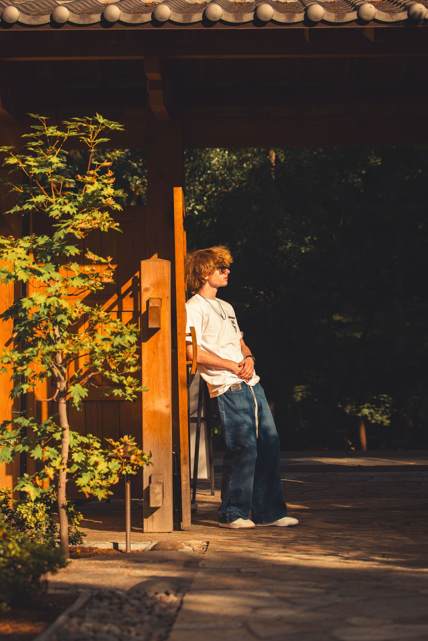 Teenage boy in white t-shirt and jeans standing under a wooden Japanese garden structure at sunset.