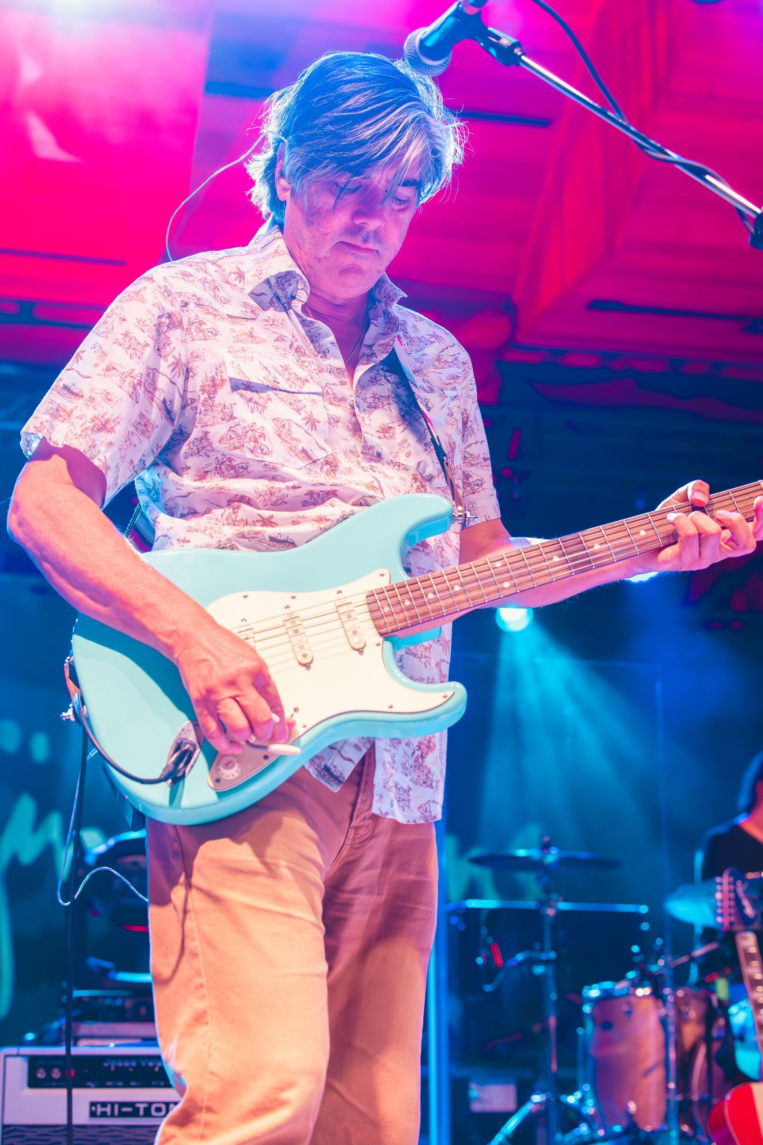 Guitarist plays a light blue Stratocaster electric guitar under colorful stage lighting.