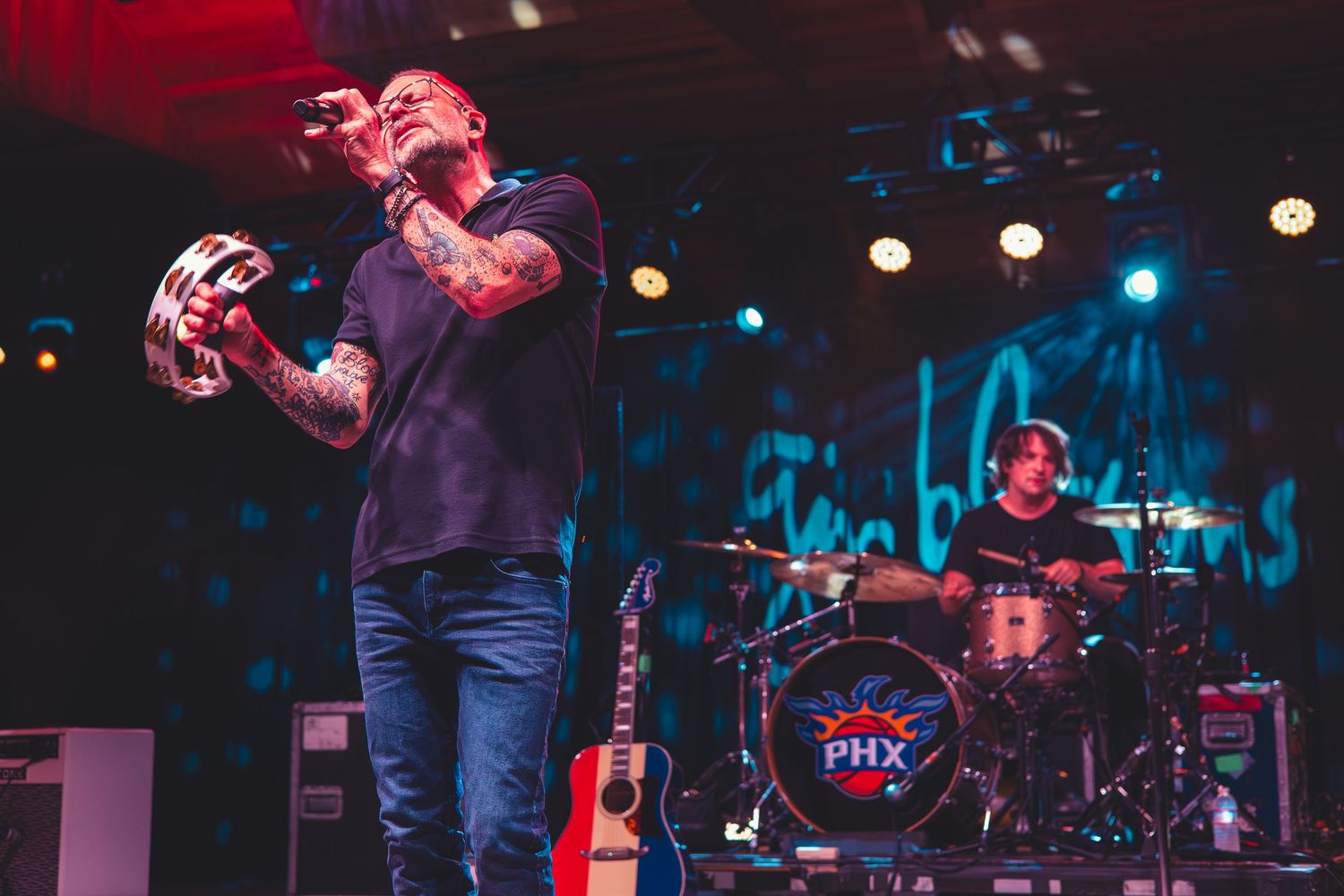 Tattooed vocalist holding a tambourine performing on stage with a drummer and acoustic guitar visible behind him.