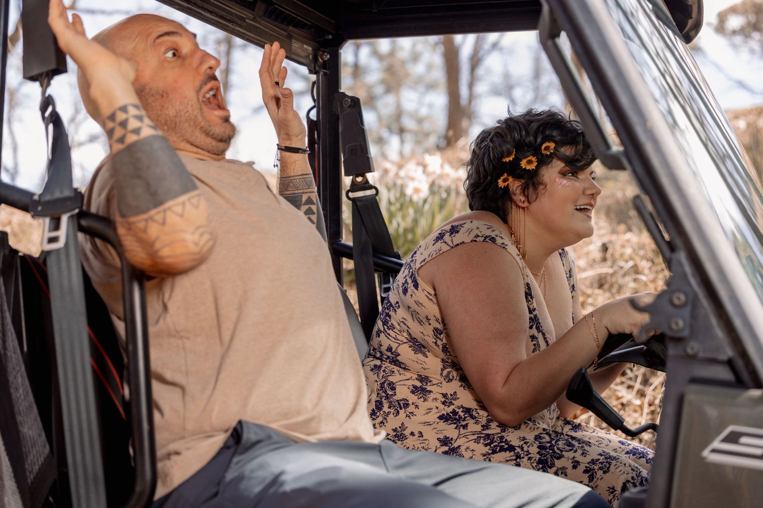 Couple laughing in UTV, man raising hands while woman drives through outdoor terrain.