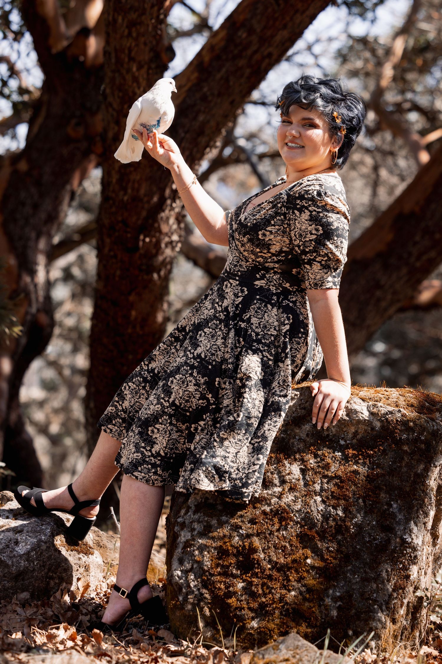 Smiling woman in vintage floral dress holds a white dove aloft while perched on a sun-dappled rock in a forest.