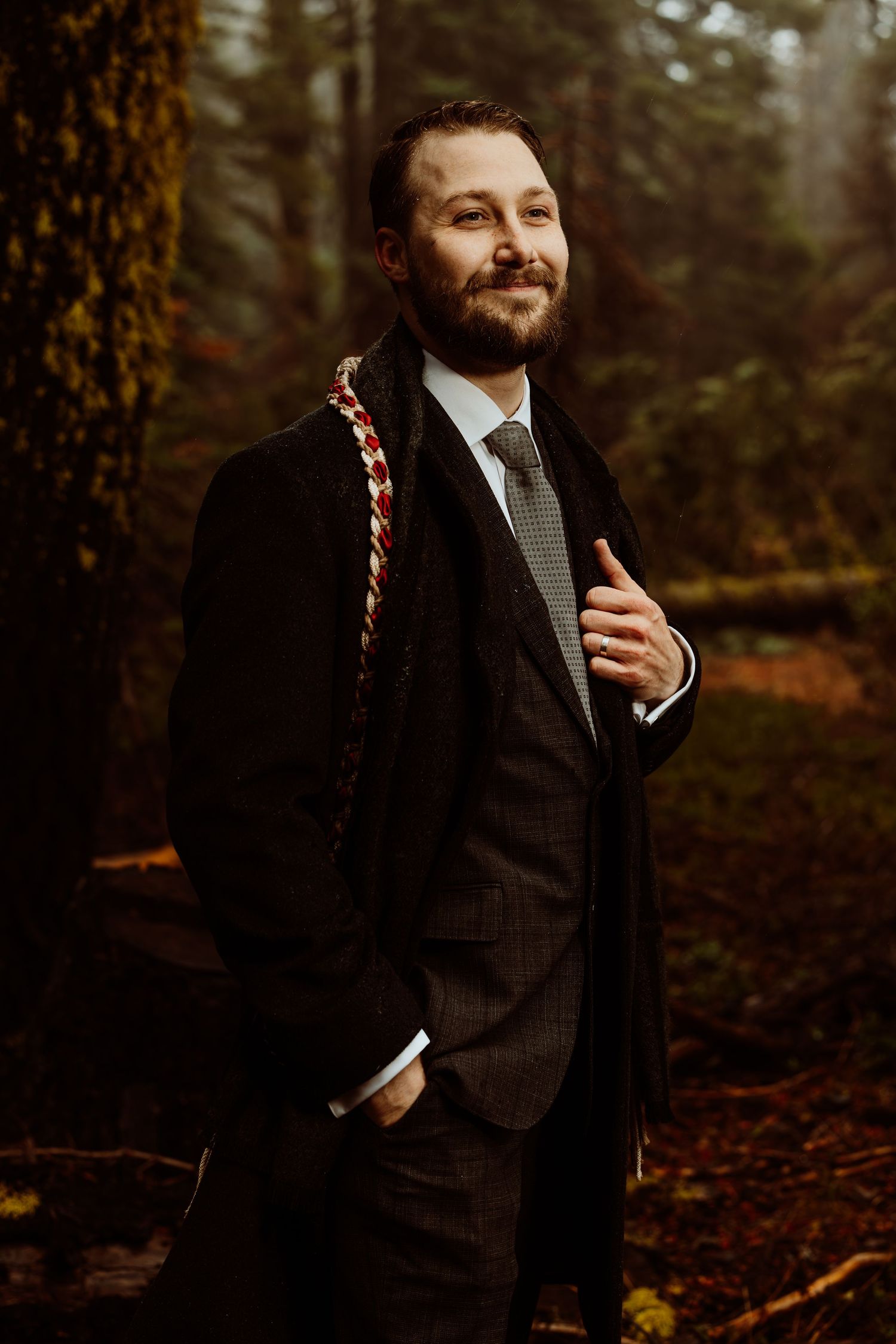 Bearded groom in dark pinstripe suit with decorative braided sash, posing confidently in autumn forest.