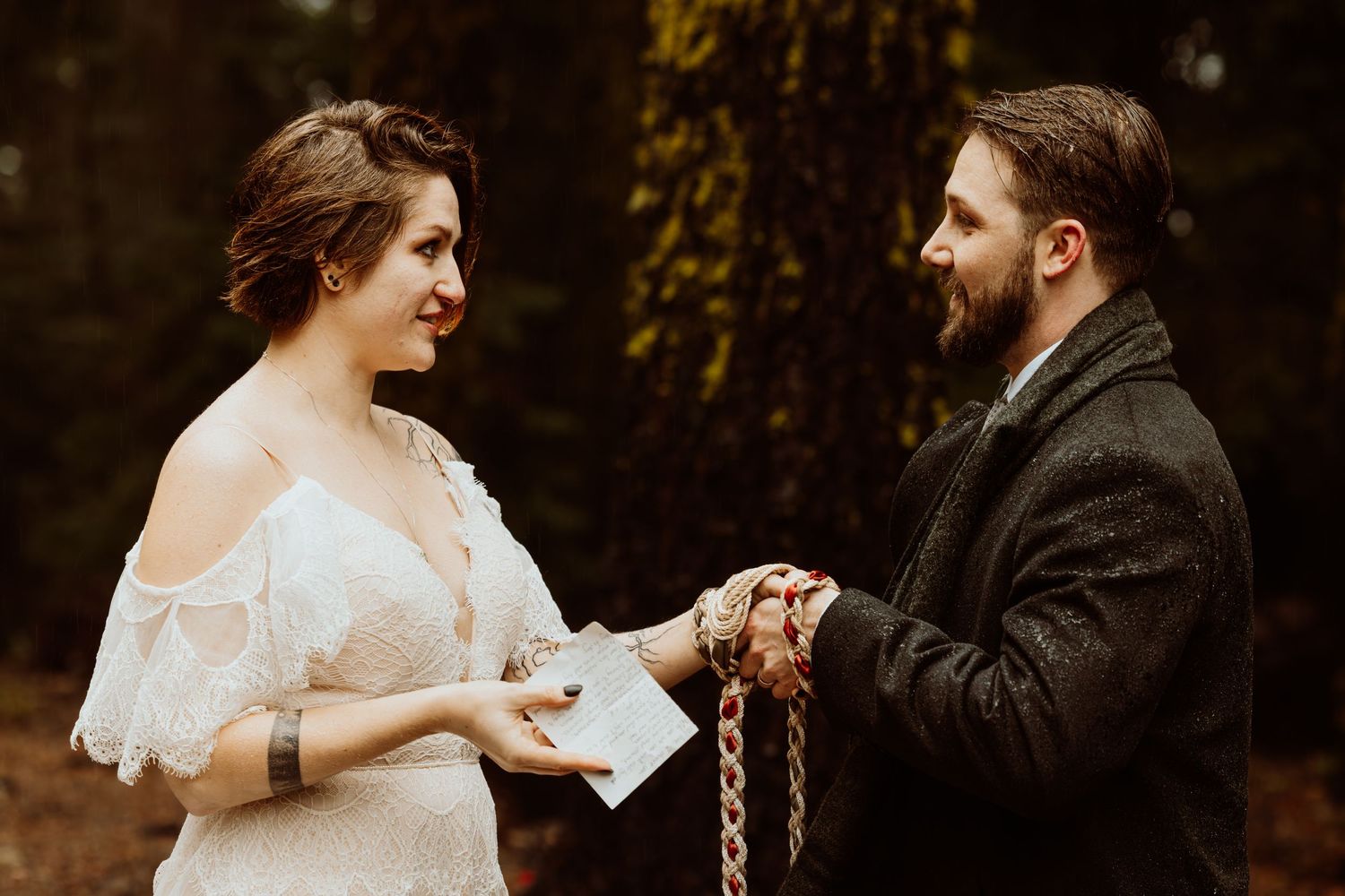 Bride reads vows from card while hands are bound in handfasting cord during forest wedding ceremony.