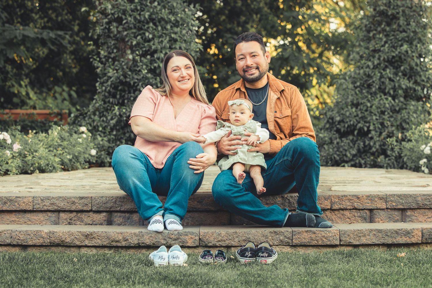 Happy young family with baby sitting on garden steps with tiny shoes displayed in front.