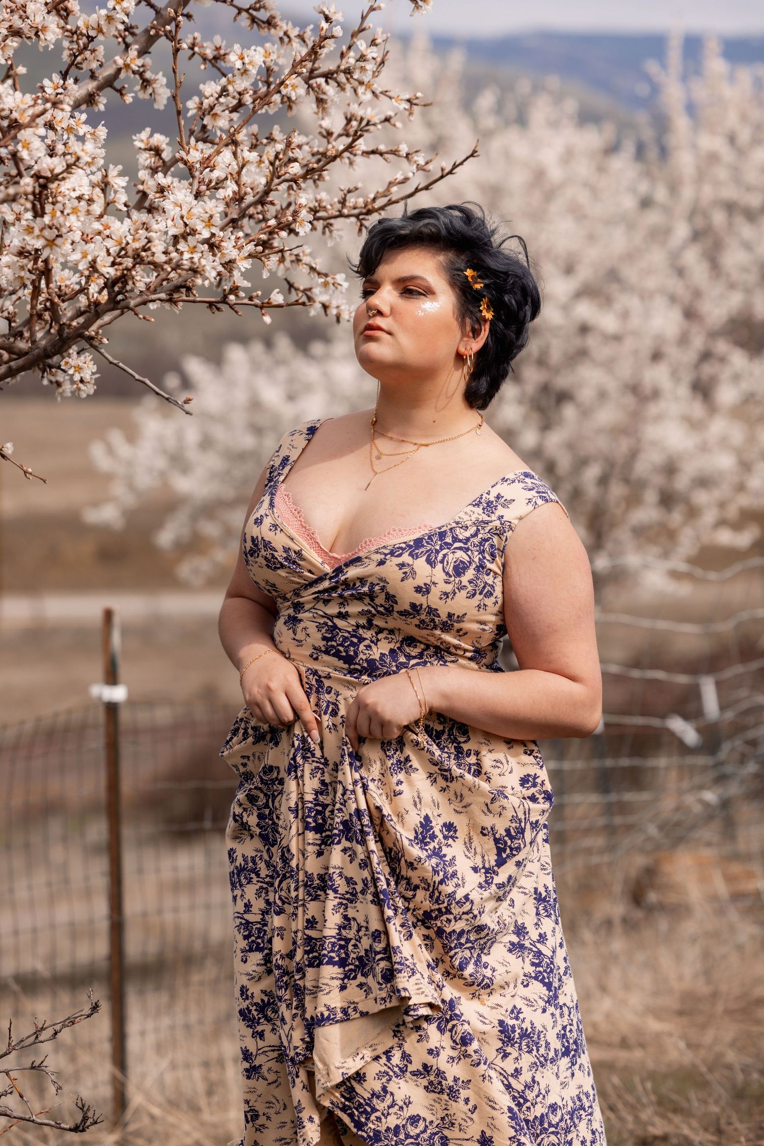Plus-size model in vintage floral dress gazing upward beside flowering spring blossoms outdoors.
