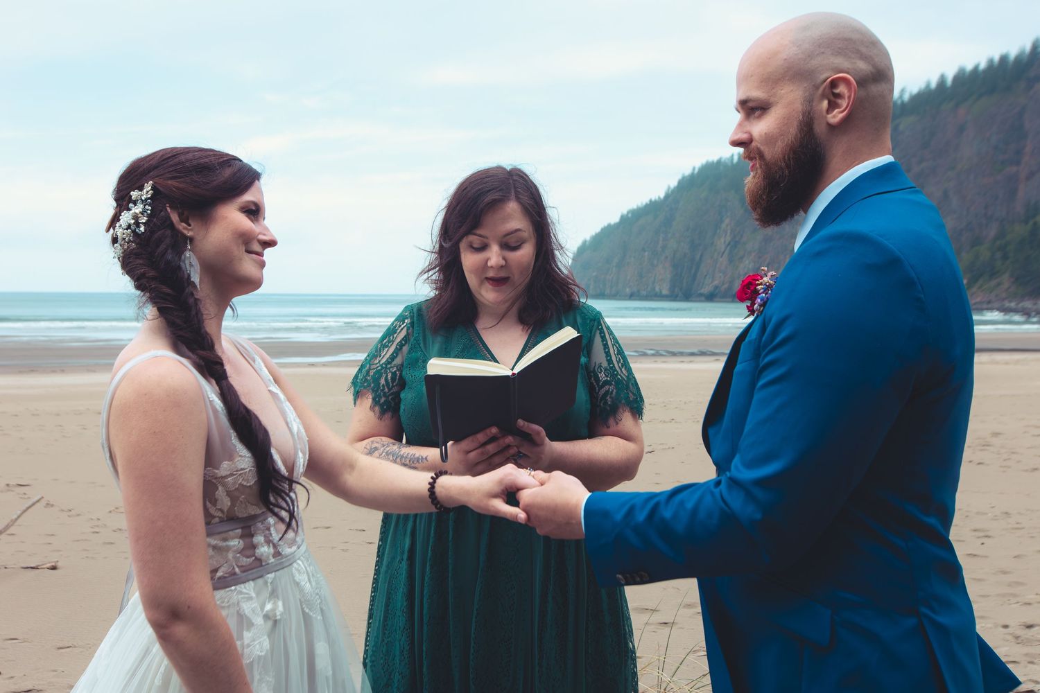 Bride and groom hold hands joyfully during beach wedding ceremony as officiant reads from a book.