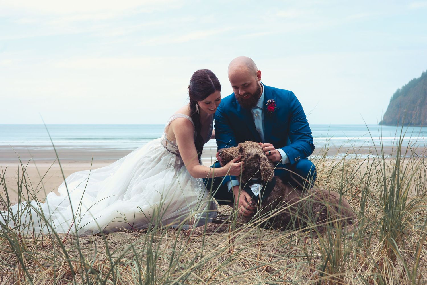 Bride in white gown and groom in blue suit pet their dog among beach grass.