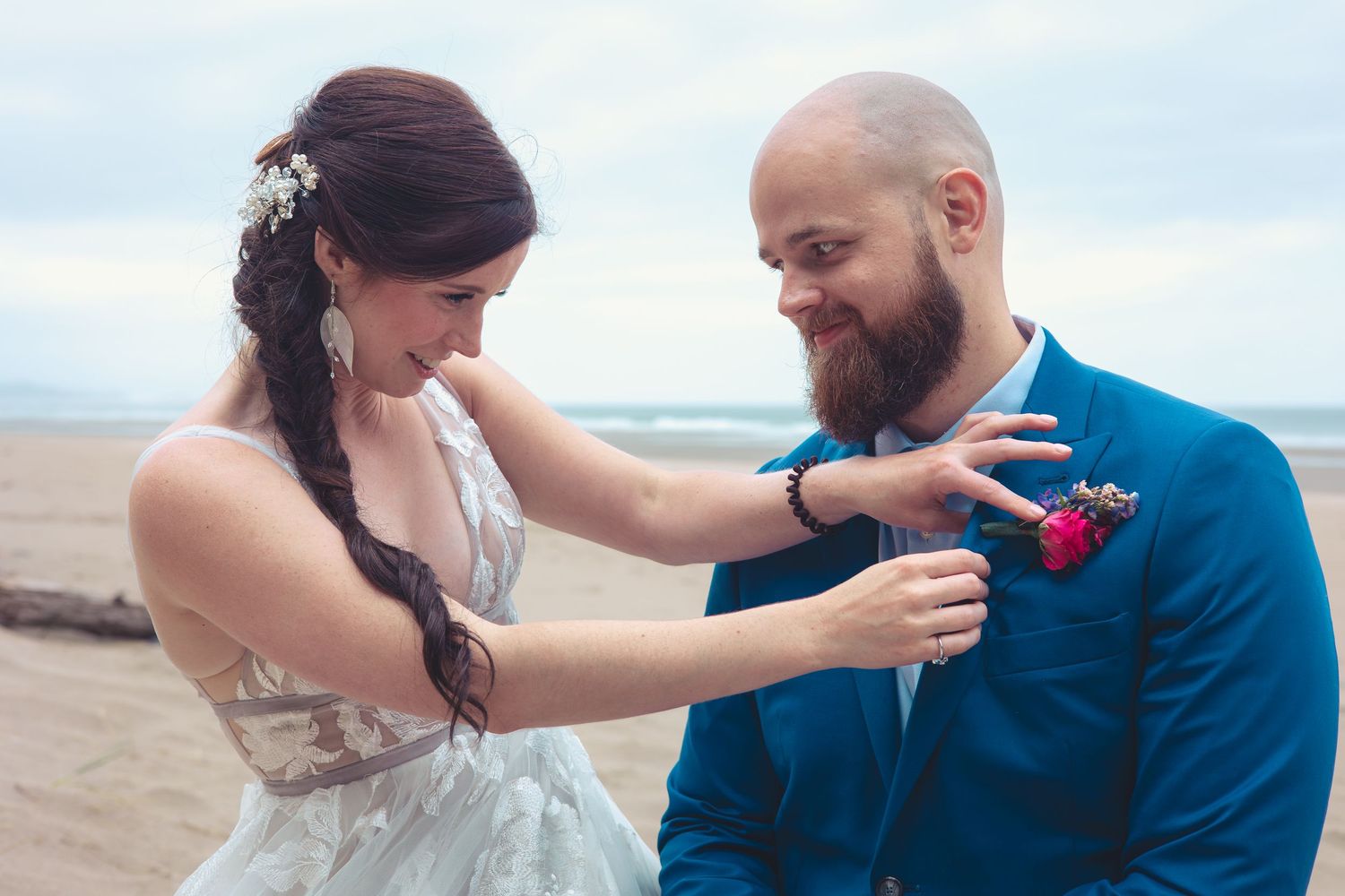 Smiling bride pins pink boutonniere on laughing groom in blue suit at beach wedding.