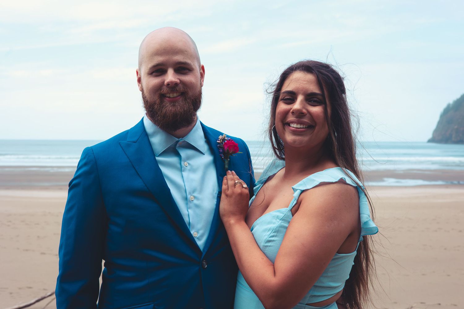 Bald groom in blue suit poses with smiling woman in light blue dress on beach.