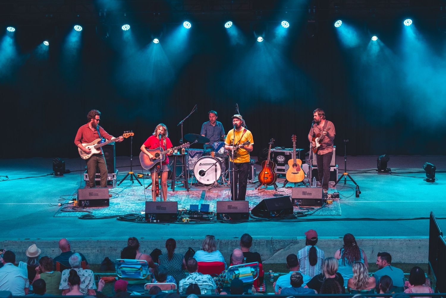 Five-piece folk band performing under teal stage lights on an indoor concert stage for a seated audience.