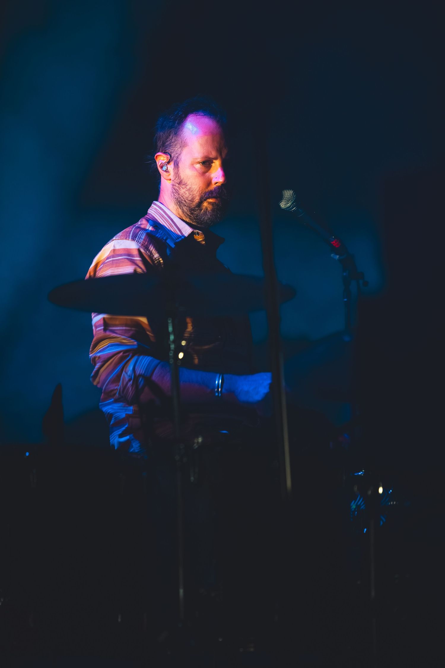 Bearded male drummer performing on stage bathed in blue and purple concert lighting with cymbal visible.