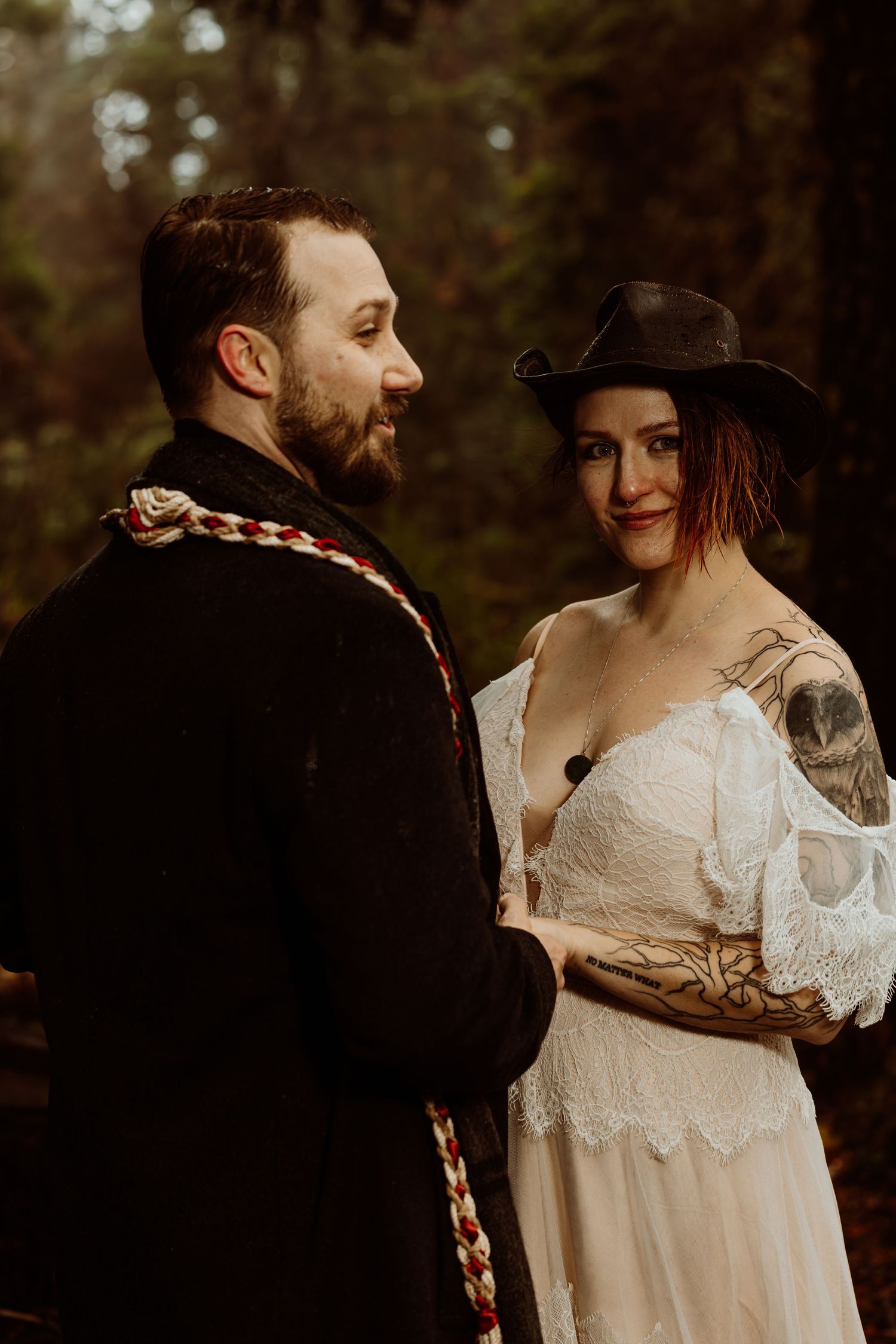 Wedding couple holds hands in dark forest, bride wearing white lace off-shoulder dress and black cowboy hat with arm tattoos.