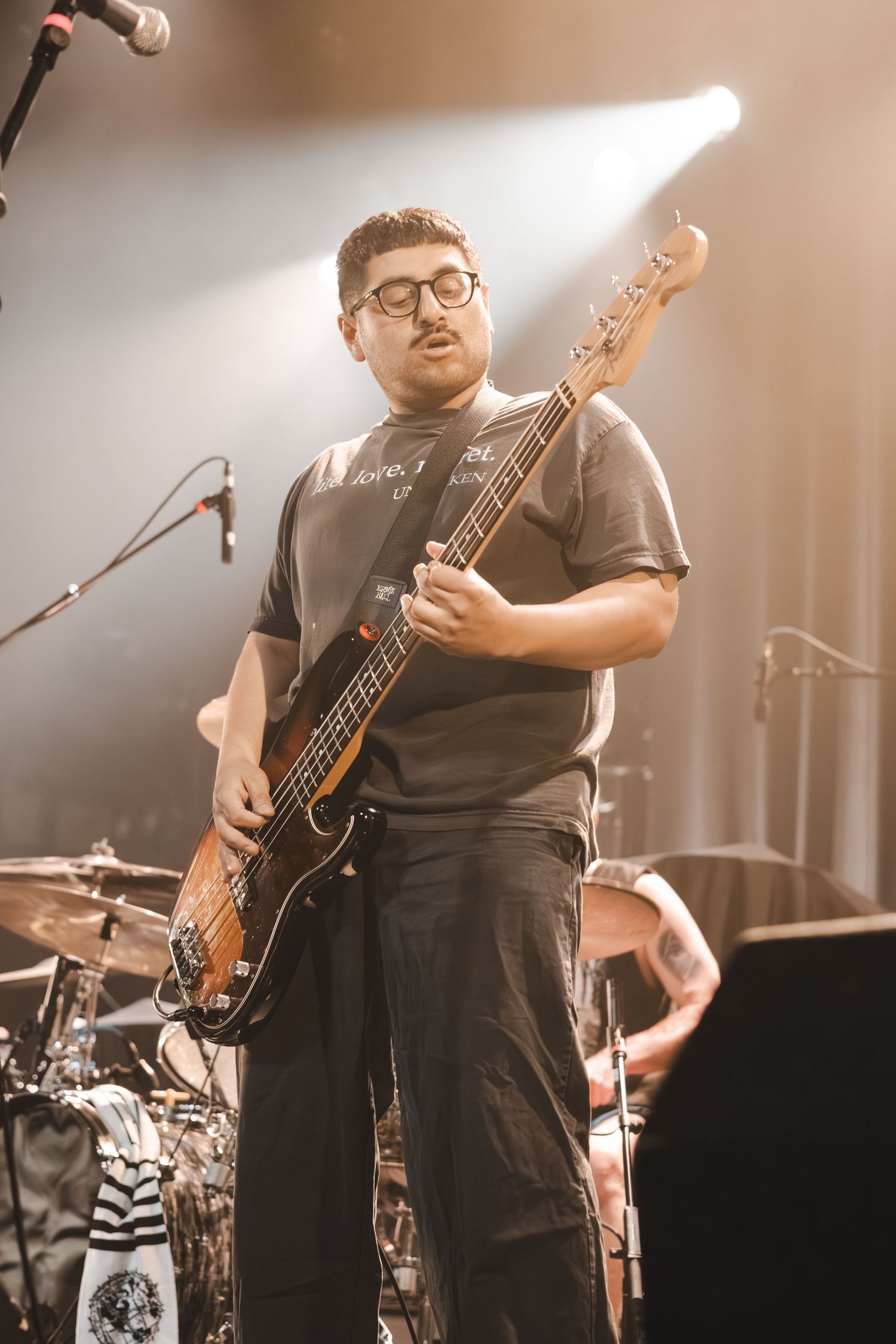Male bassist with glasses plays electric bass guitar under warm golden stage spotlight.