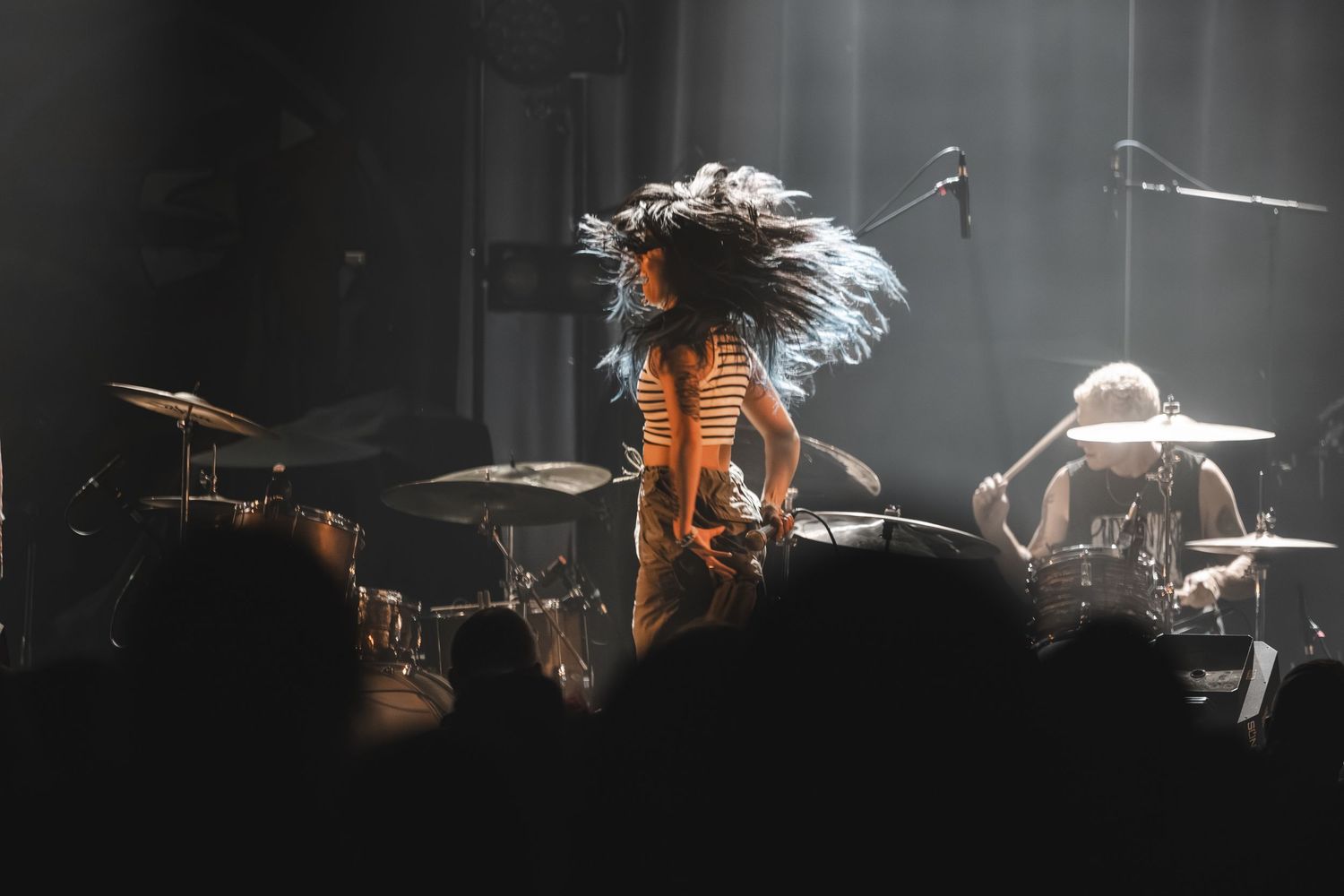 Color concert photo of performer with wild flying hair on stage under dramatic spotlights with drummer behind.