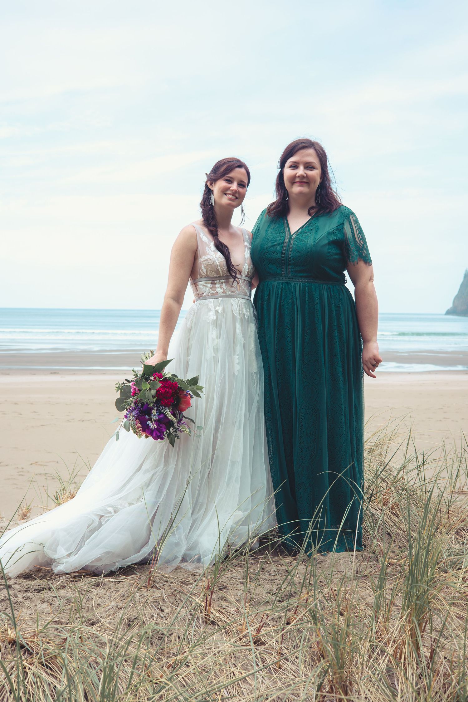 A bride in a white lace gown and woman in an emerald green dress pose on a beach with bouquet.