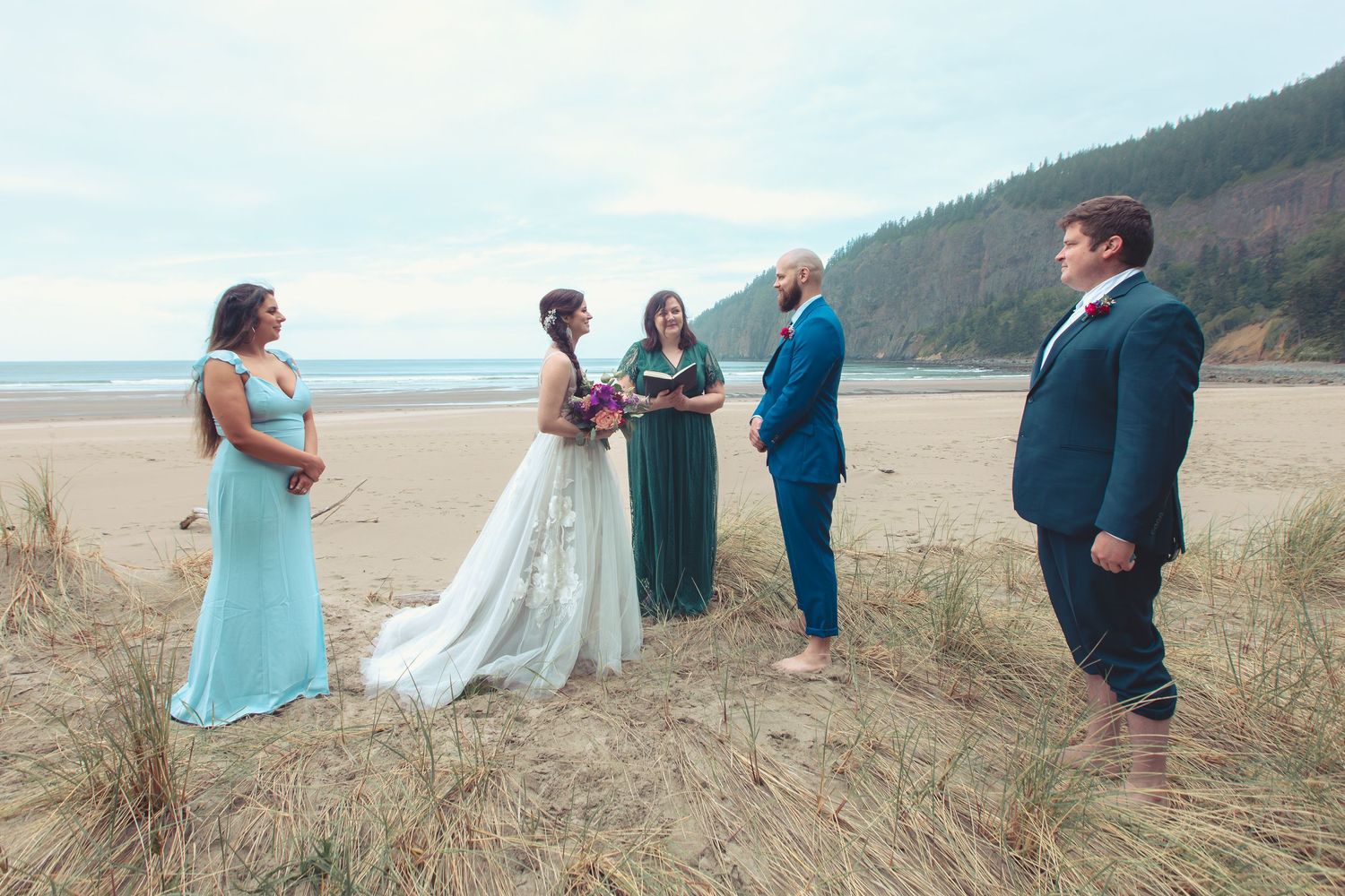Wedding party stands on a beach with grassy dunes, as officiant reads vows to bride and groom.