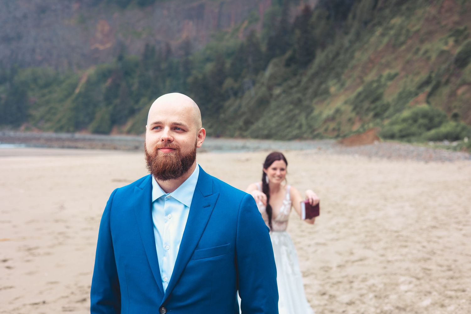 Groom in blue suit stands on beach while bride in white dress approaches smiling.