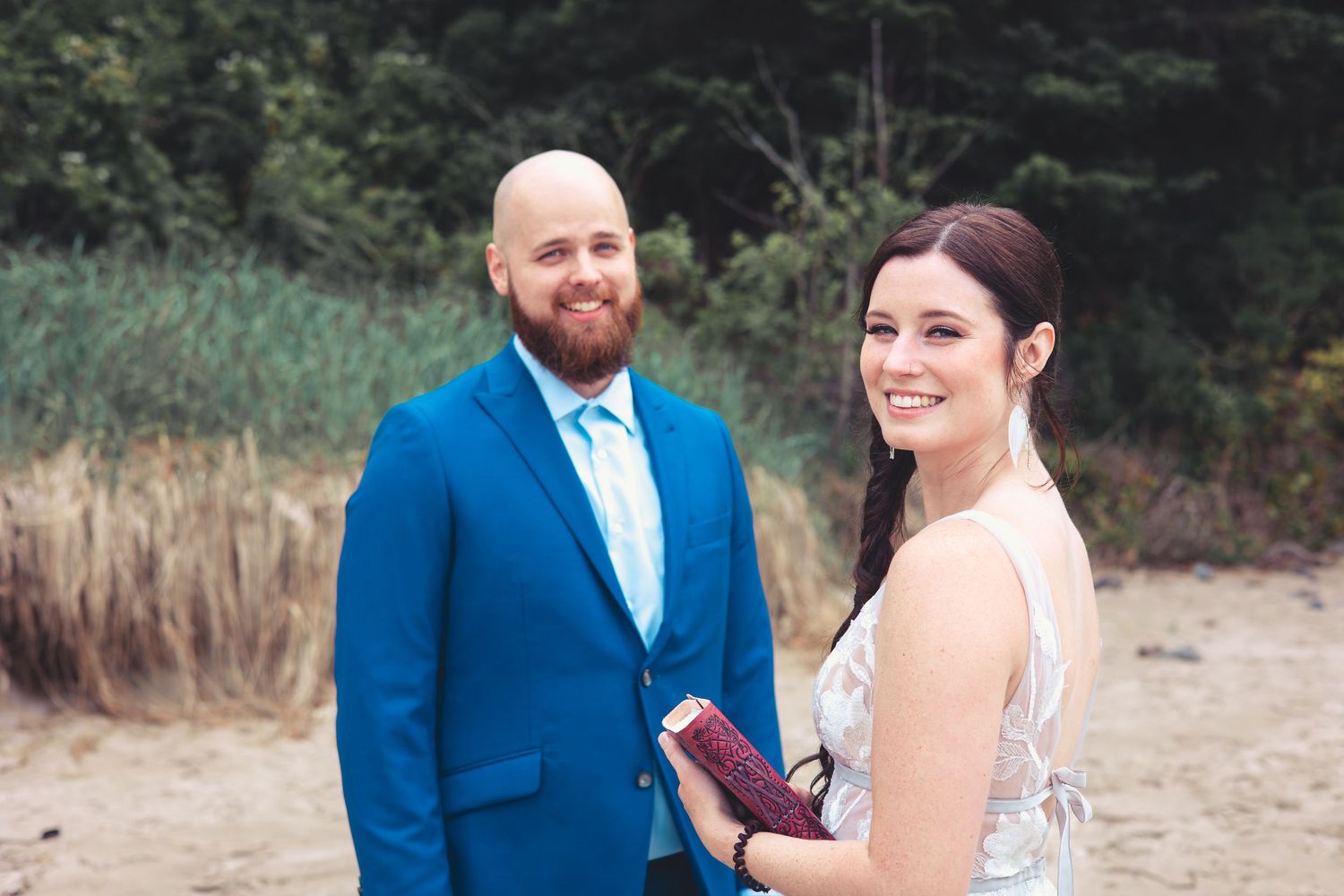 Bride holding a book smiles at camera while groom in blue suit stands beside her.
