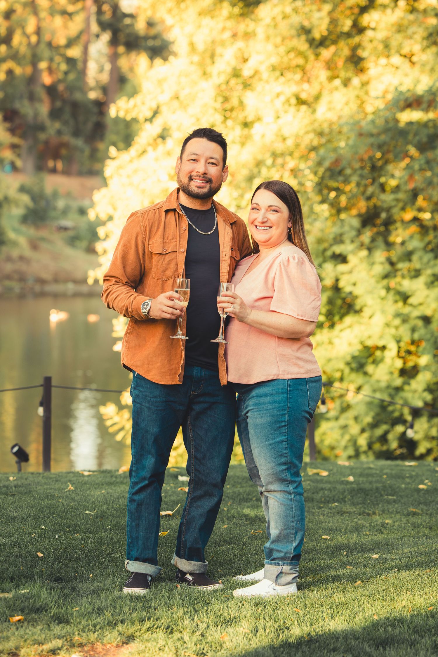 Happy couple holding champagne flutes standing together on grass near a tranquil autumn pond.